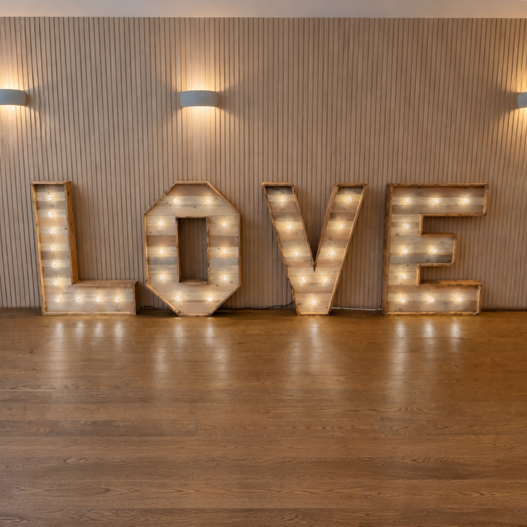 Decorative wooden letters spelling 'LOVE' with illuminated bulbs, placed against a light wooden paneled wall with warm wall sconces.