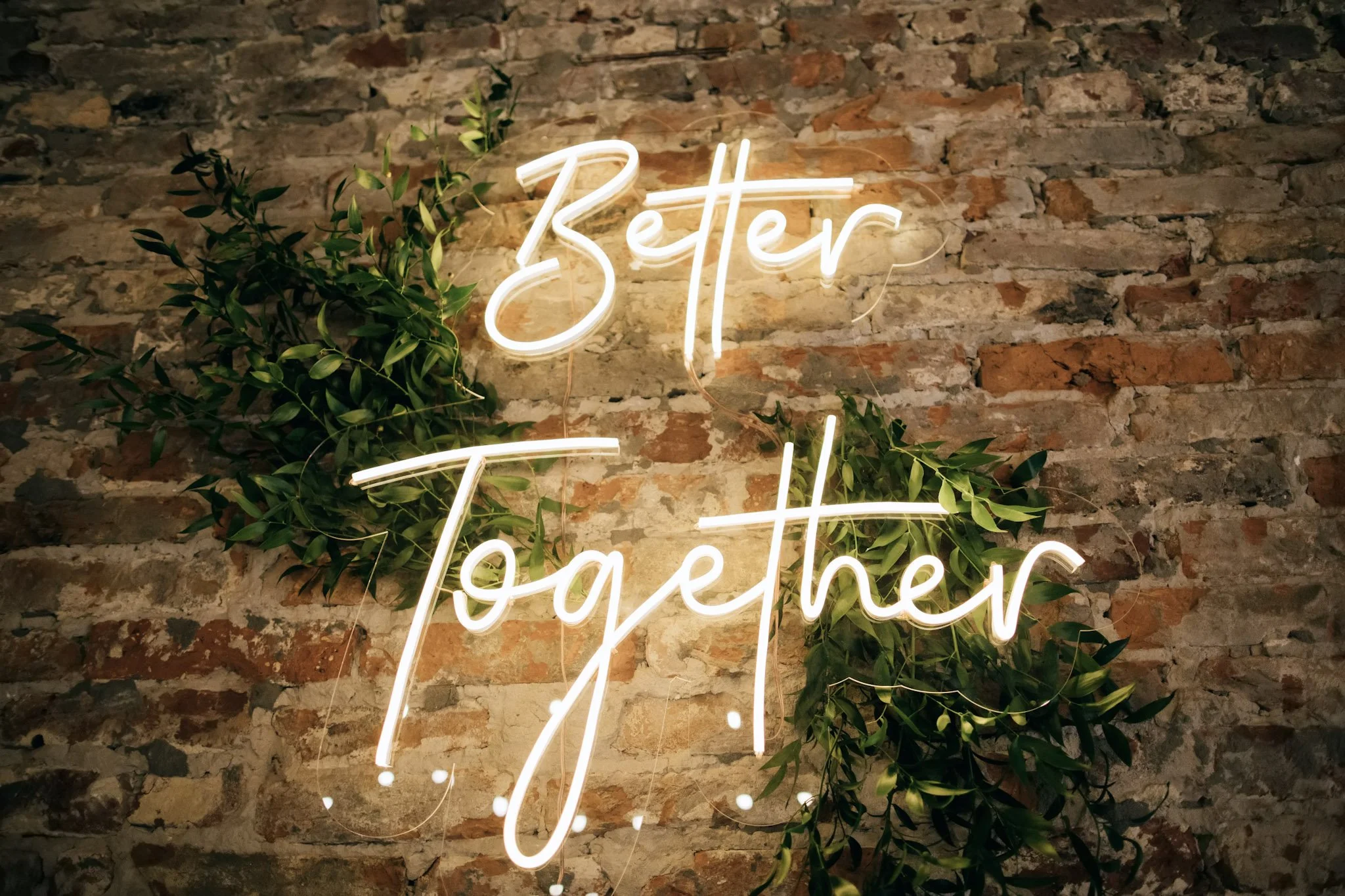 Neon sign on a brick wall that says 'Better Together' with some green leafy plants nearby.