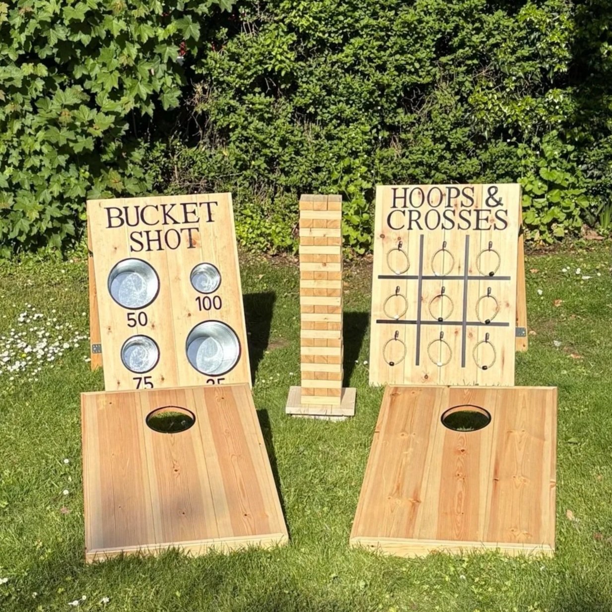 Two wooden outdoor games with painted text and graphics, one labeled 'Bucket Shot' with holes and point values, and the other labeled 'Hoops & Crosses' with a tic-tac-toe grid and metal rings, set on grass with greenery in the background.