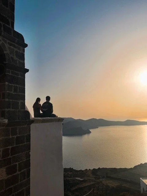 Two people sitting on a ledge overlooking a large body of water and distant mountains during sunset.