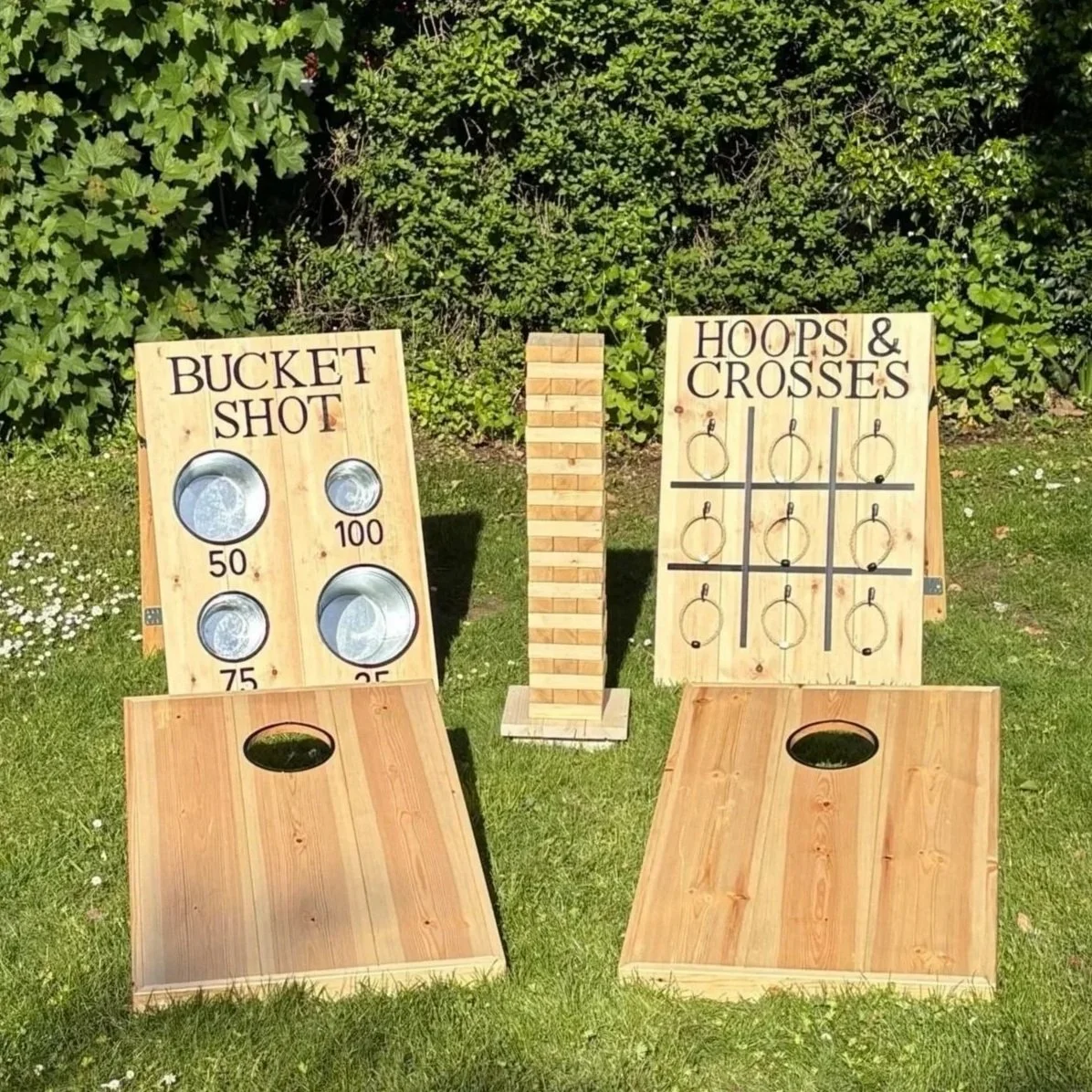 Outdoor cornhole game setup with two boards, one labeled "Bucket Shot" with scoring circles and the other labeled "Hoops & Crosses" with ring toss rings, set on a grassy area with trees in the background.