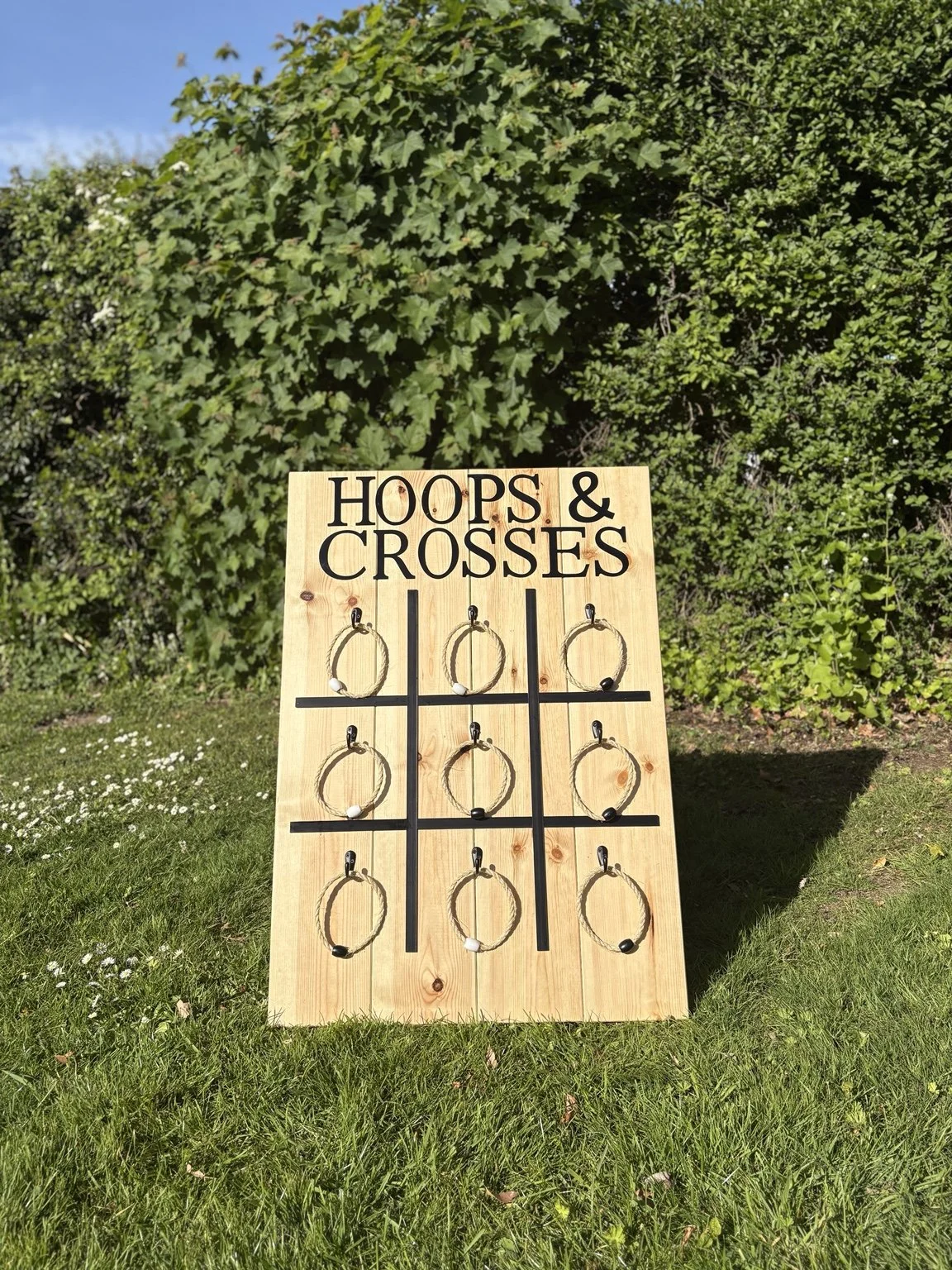 A wooden Tic Tac Toe game board with handmade hoops for a game called 'Hoops & Crosses', set outdoors on grass with bushes and a blue sky in the background.