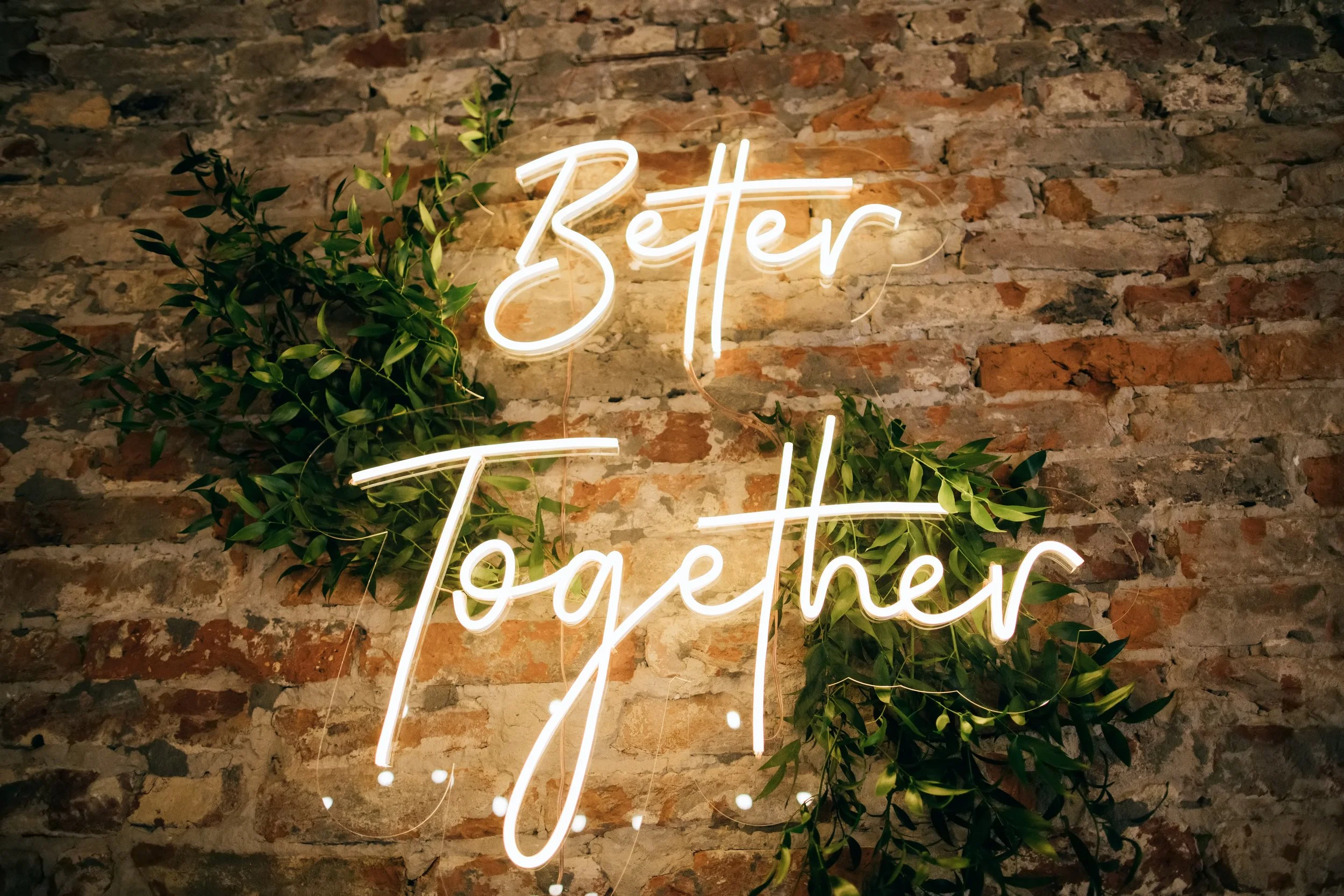 Neon sign spelling "Better Together" mounted on a brick wall, with green leafy plants surrounding it.