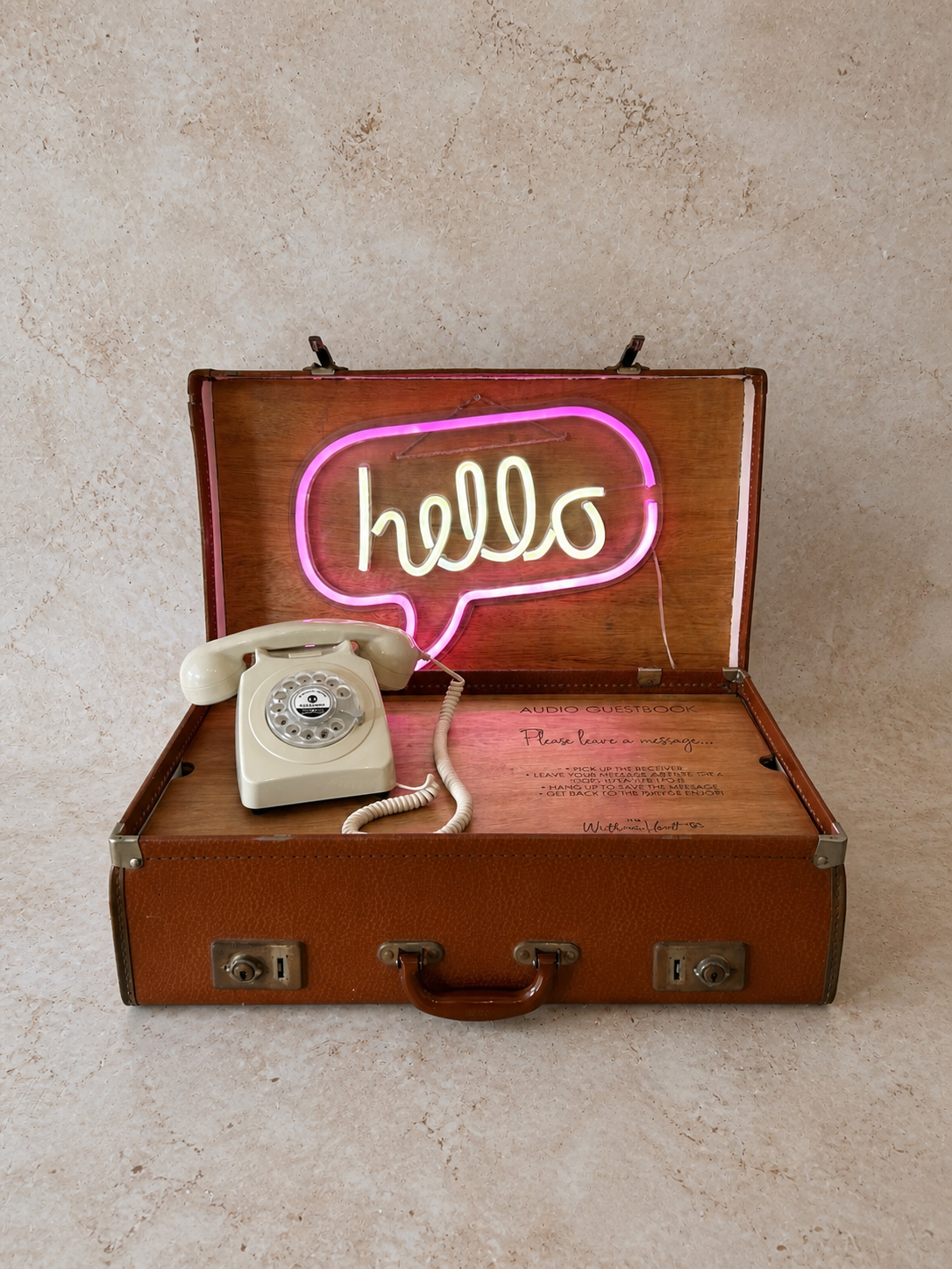 Open vintage brown suitcase with a neon sign inside that says 'hello' in cursive, and a beige rotary telephone resting inside the suitcase on the left.