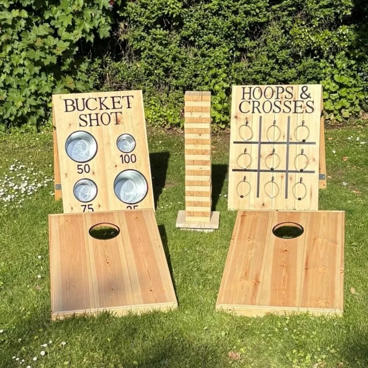 Two wooden yard games boards, one labeled "Bucket Shot" with four metal buckets marked with point values 50, 75, 100, and 25, and another labeled "Hoops & Crosses" with a tic-tac-toe grid and six hoops hanging from hooks, placed outdoors on grass with greenery in the background.