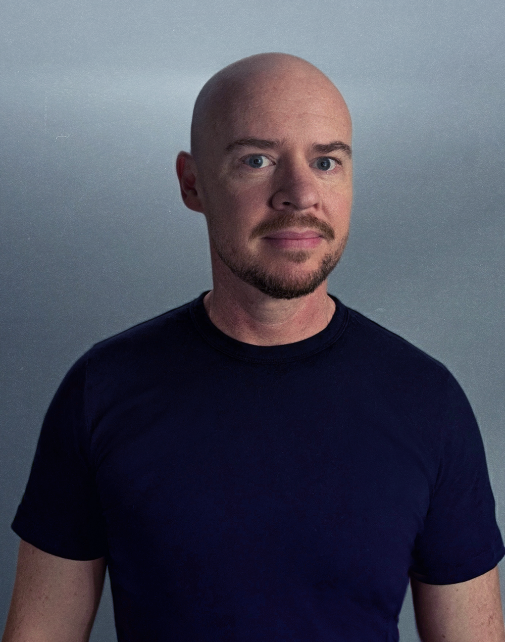Profile image of Ben Hopkinson with a shaved head, light blue eyes, and a stubble, wearing a navy t-shirt, standing against a plain, muted background.
