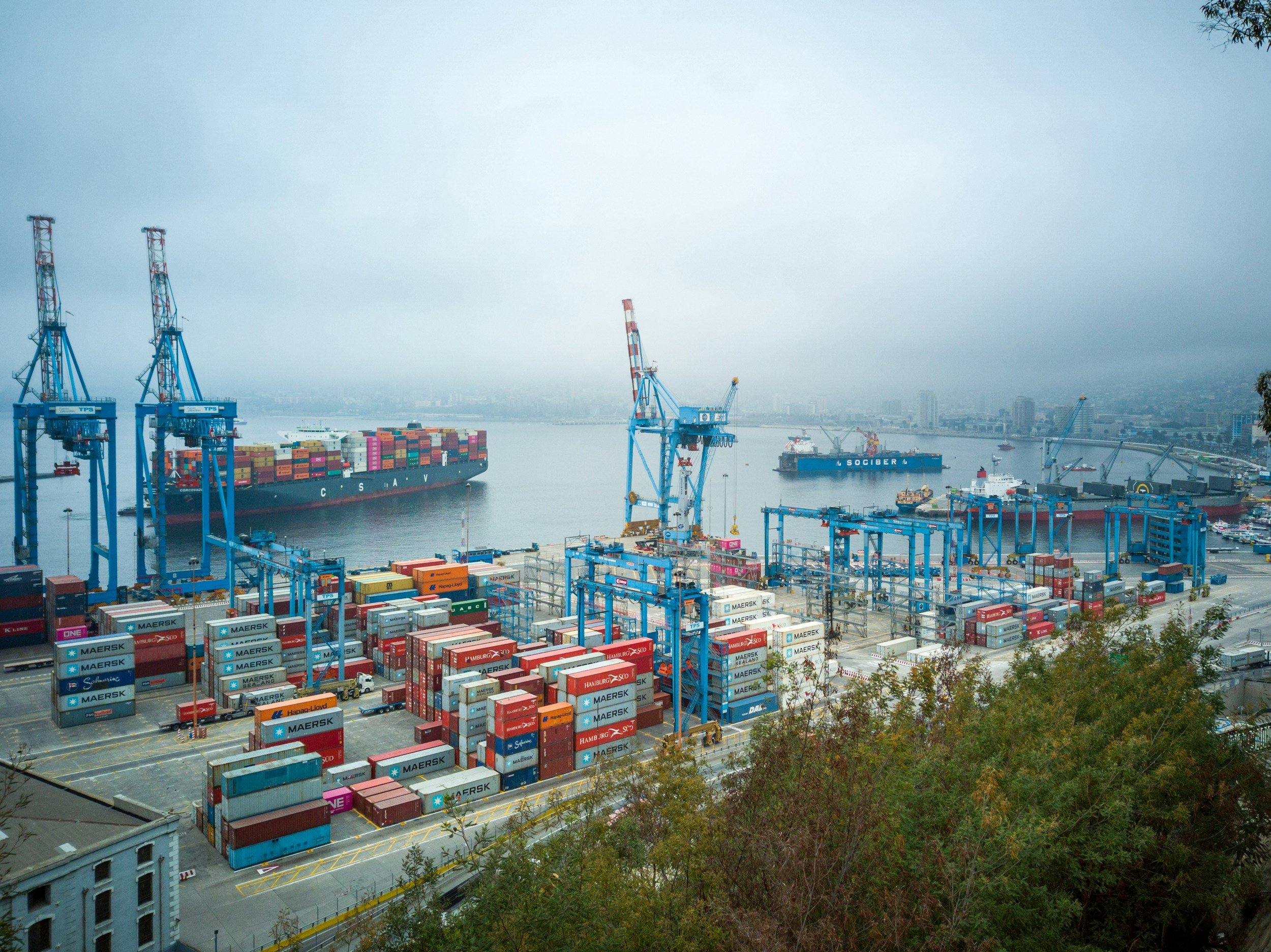 Vue d'un port rempli de conteneurs colorés et de grues de chargement, avec une mer et des bâtiments à l'horizon, sous un ciel brumeux.