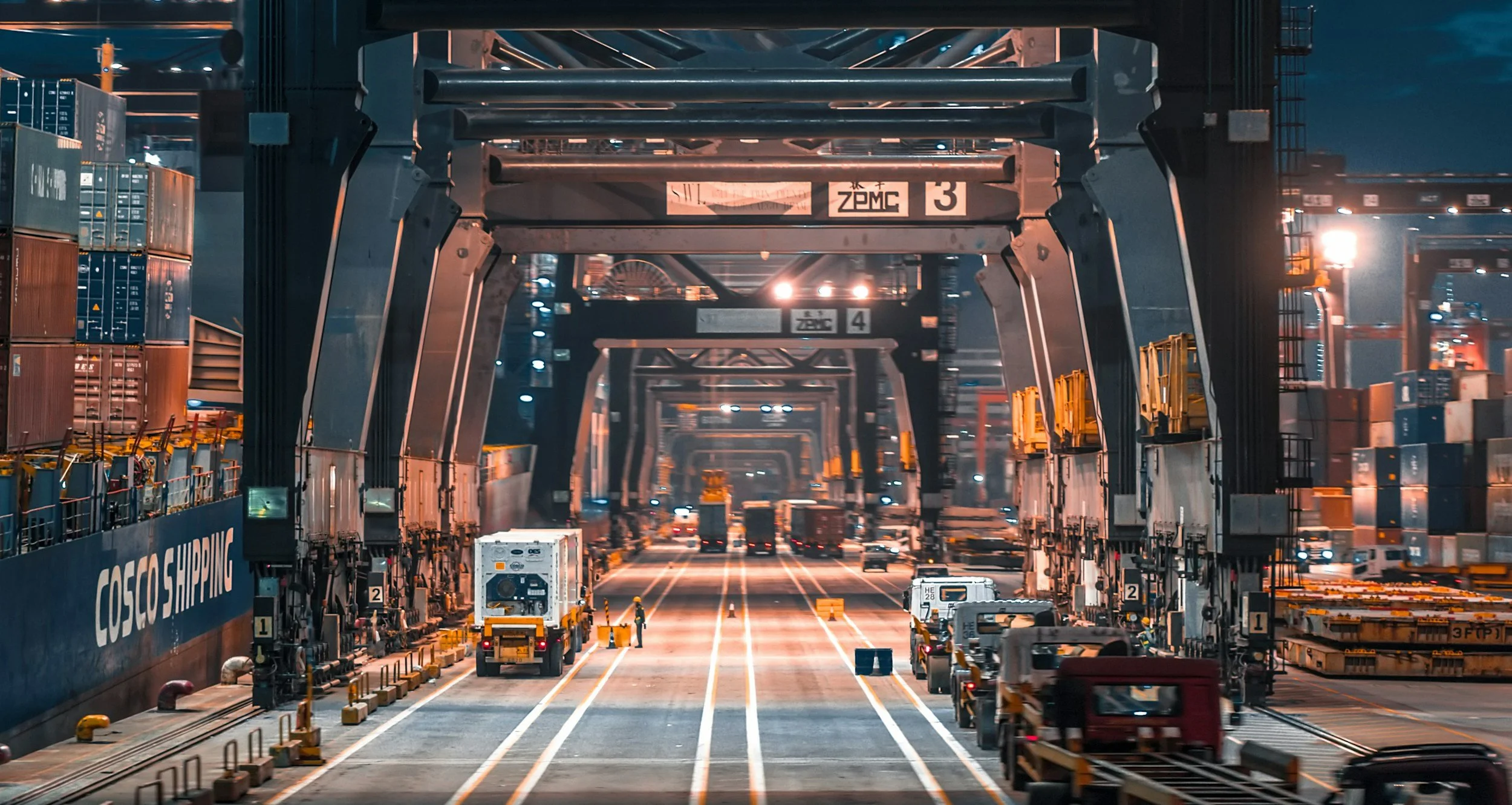 Un port industriel avec des grues de manutention, des conteneurs emballés, et des véhicules en attente sous un éclairage nocturne, illustrant une zone de chargement et de déchargement maritime.