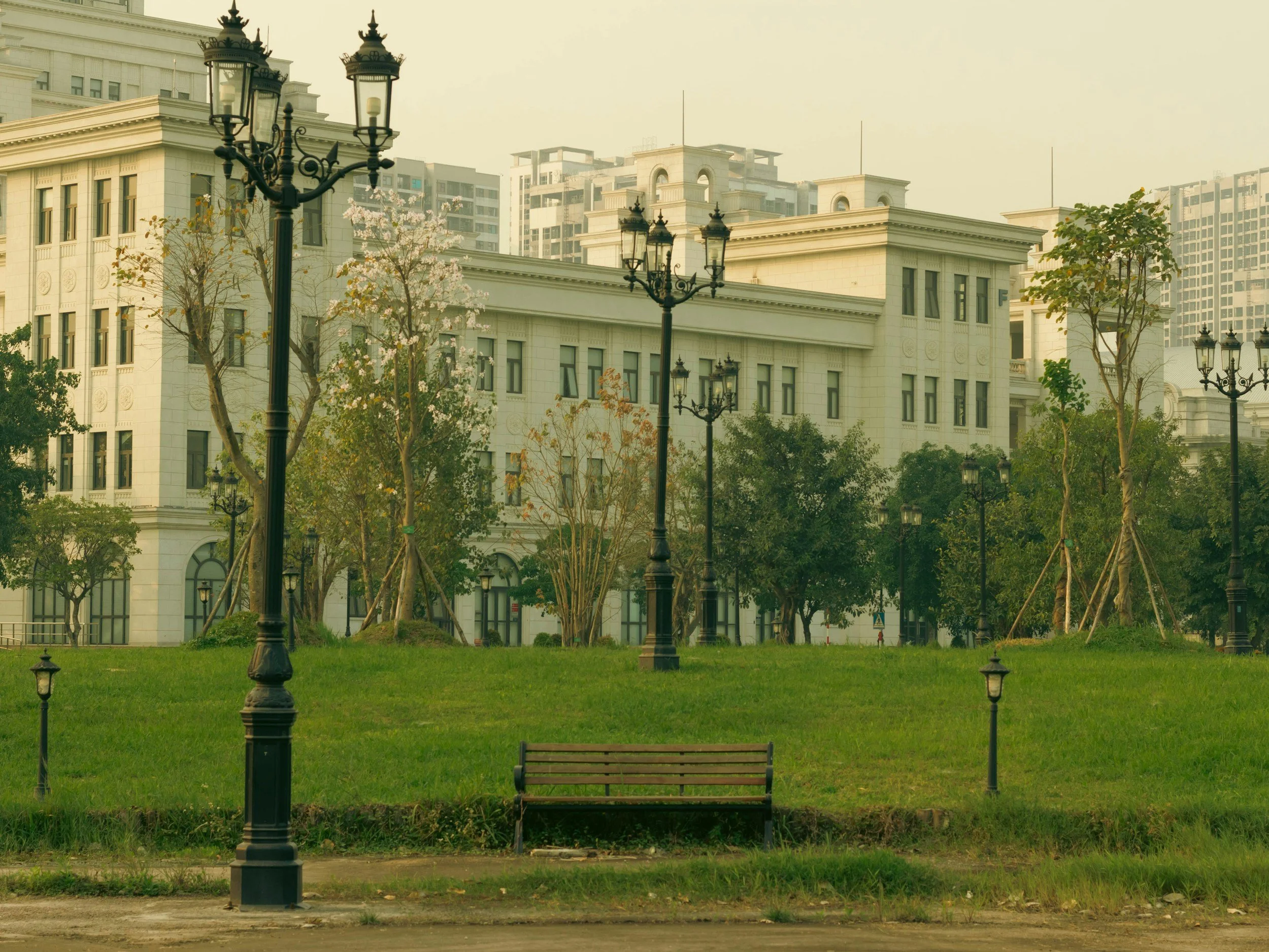 Un parc avec un banc et des lampadaires en fer noir, entouré de quelques arbres en fleurs avec des immeubles modernes en arrière-plan.