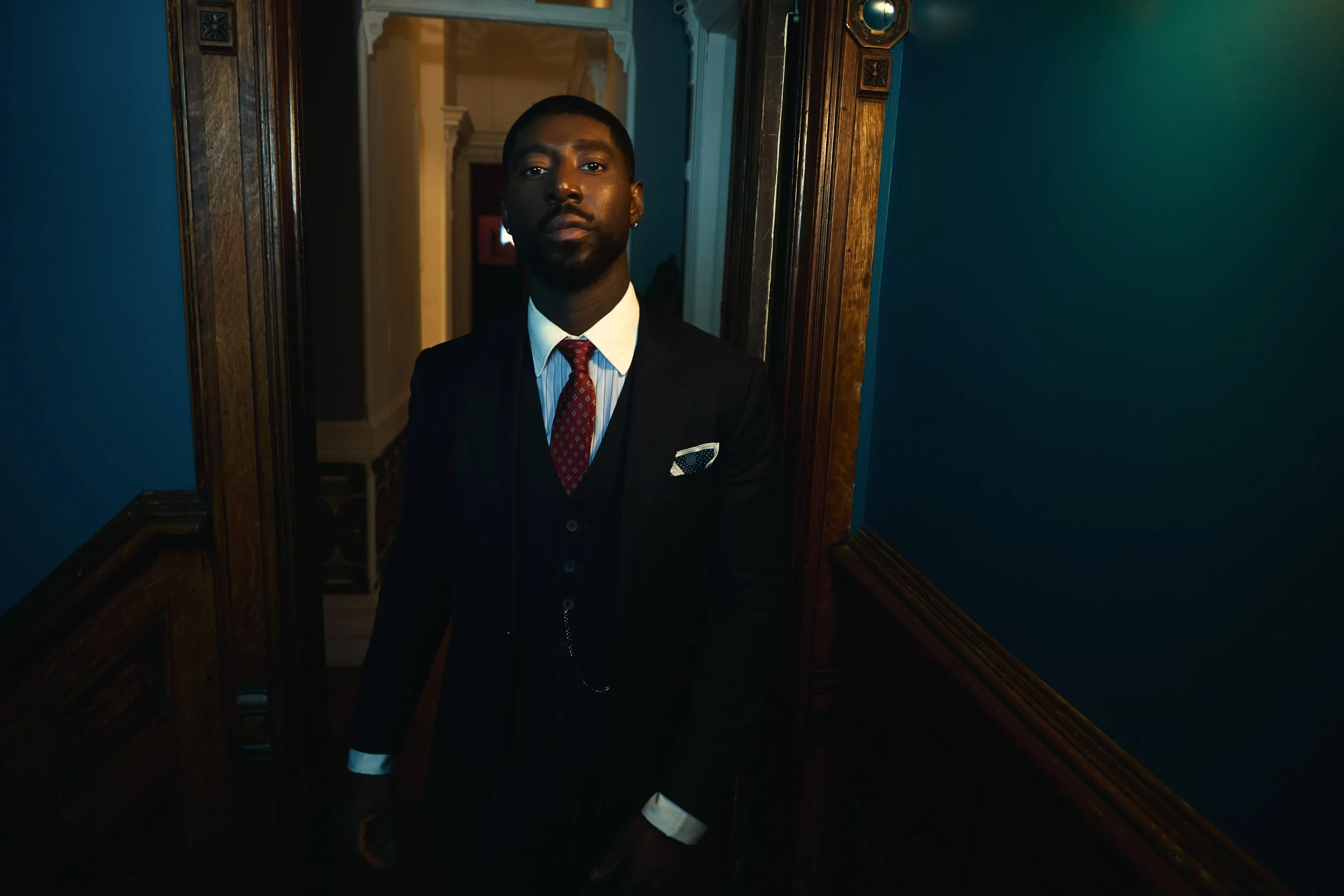 A young man in a black suit with a white shirt, red patterned tie, and pocket square standing in front of a mirror in a dimly lit room with blue walls and wooden trim.