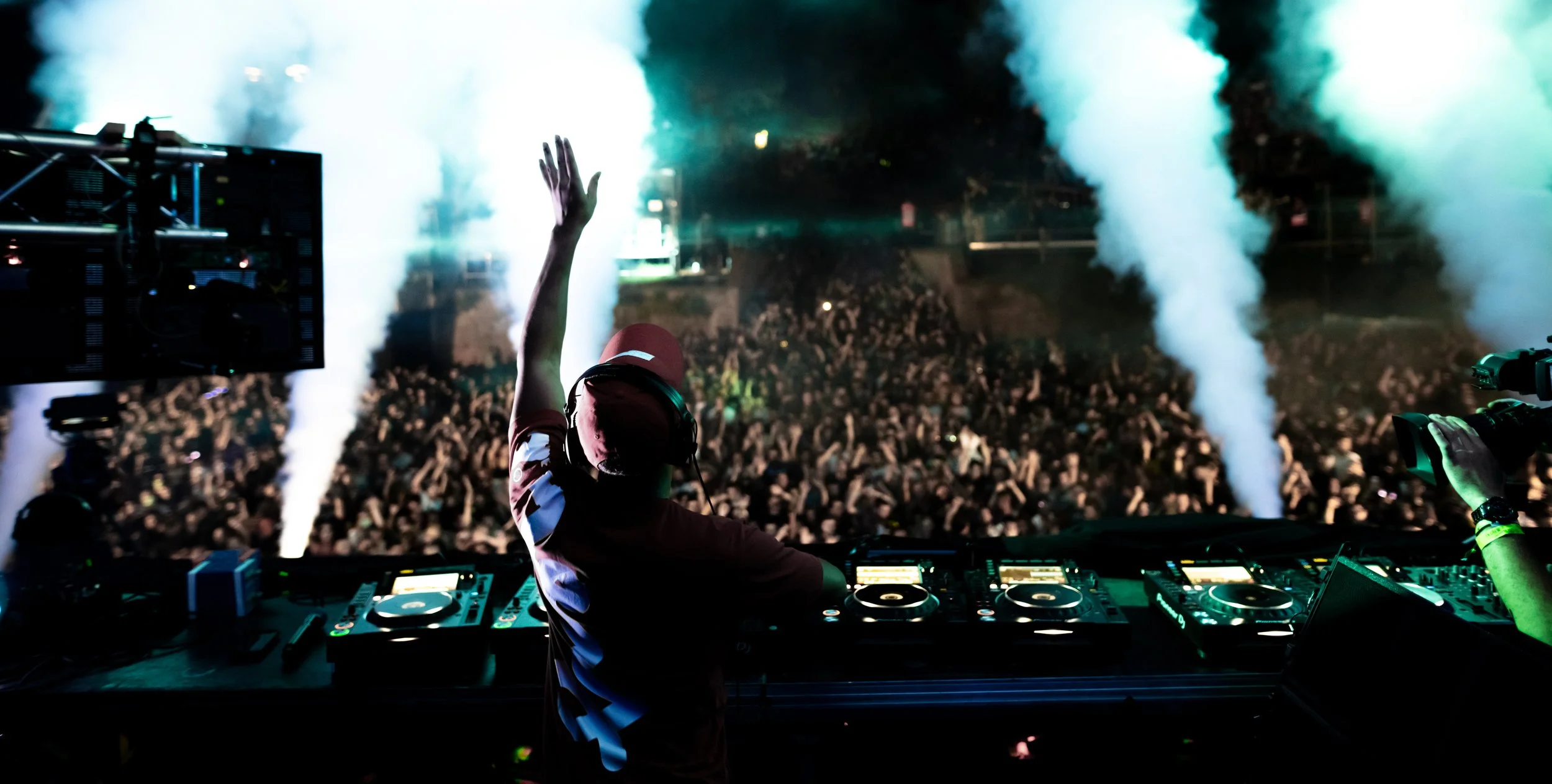 DJ performing at a large night-time music festival with a crowd, smoke effects, and stage lighting in the background.