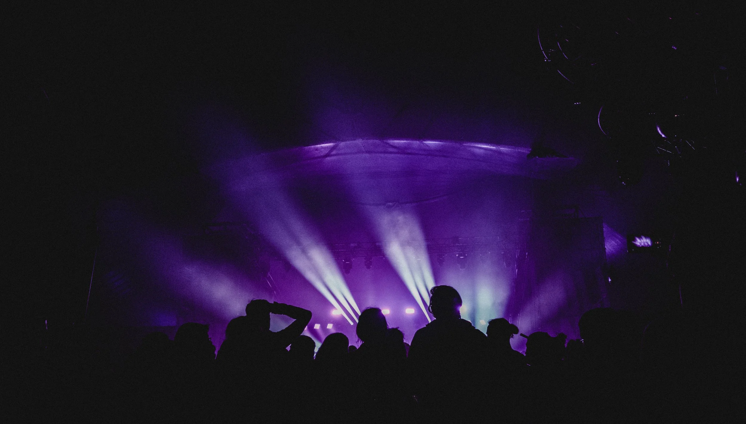 Dark concert scene with purple stage lights, silhouettes of audience members, and beams of light projecting upwards.