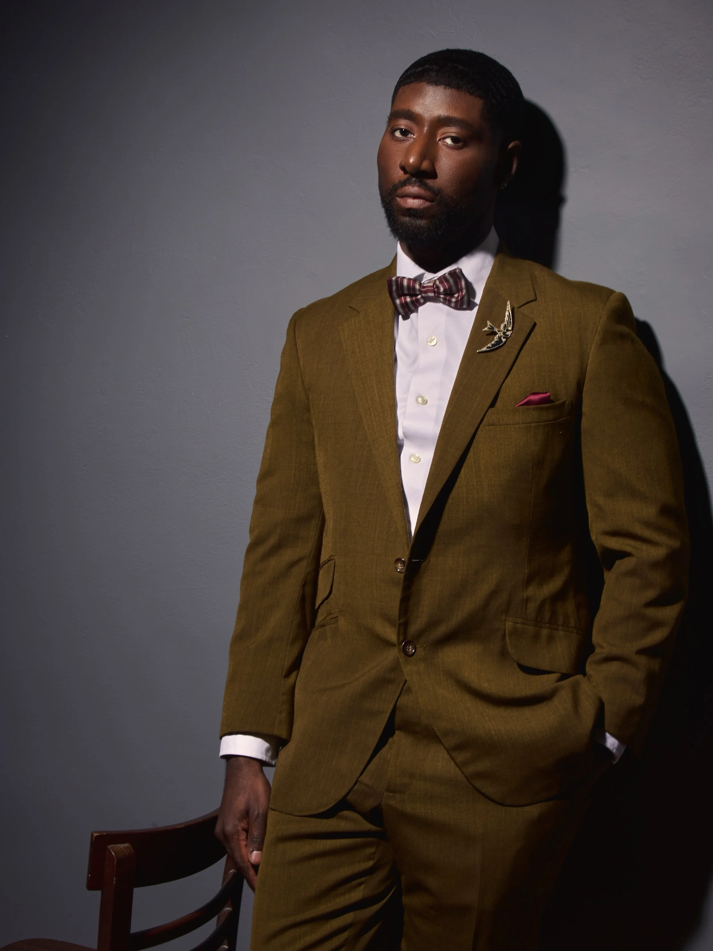 A man in a brown suit with a bow tie, standing next to a wooden chair against a gray wall.