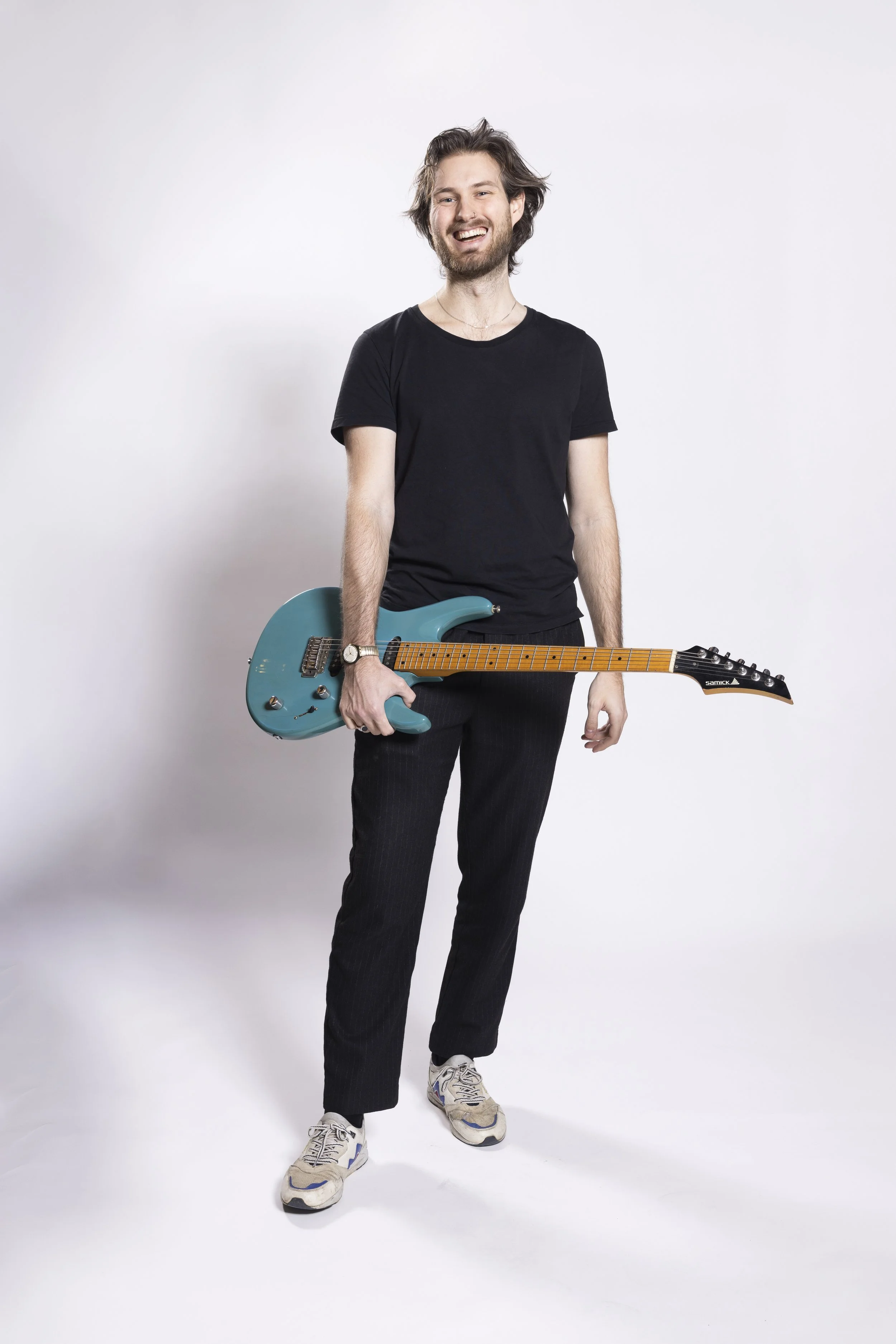 Ein lachender Mann mit einem blauen E-Gitarre in der Hand, trägt ein schwarzes T-Shirt, schwarze Hose und Sportschuhe, steht vor weißem Hintergrund.