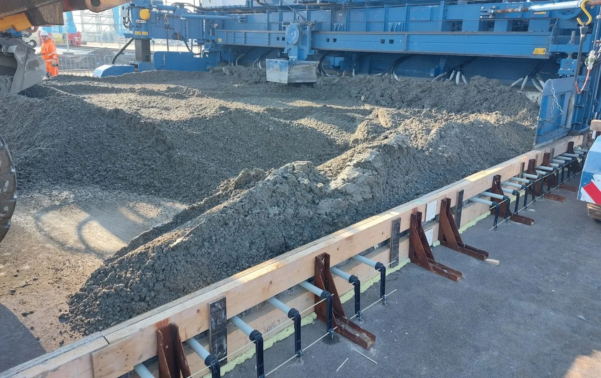 Een bouwplaats waar beton wordt gestort in een houten mal, omgeven door bouwmateriaal en werkers in veiligheidskleding.