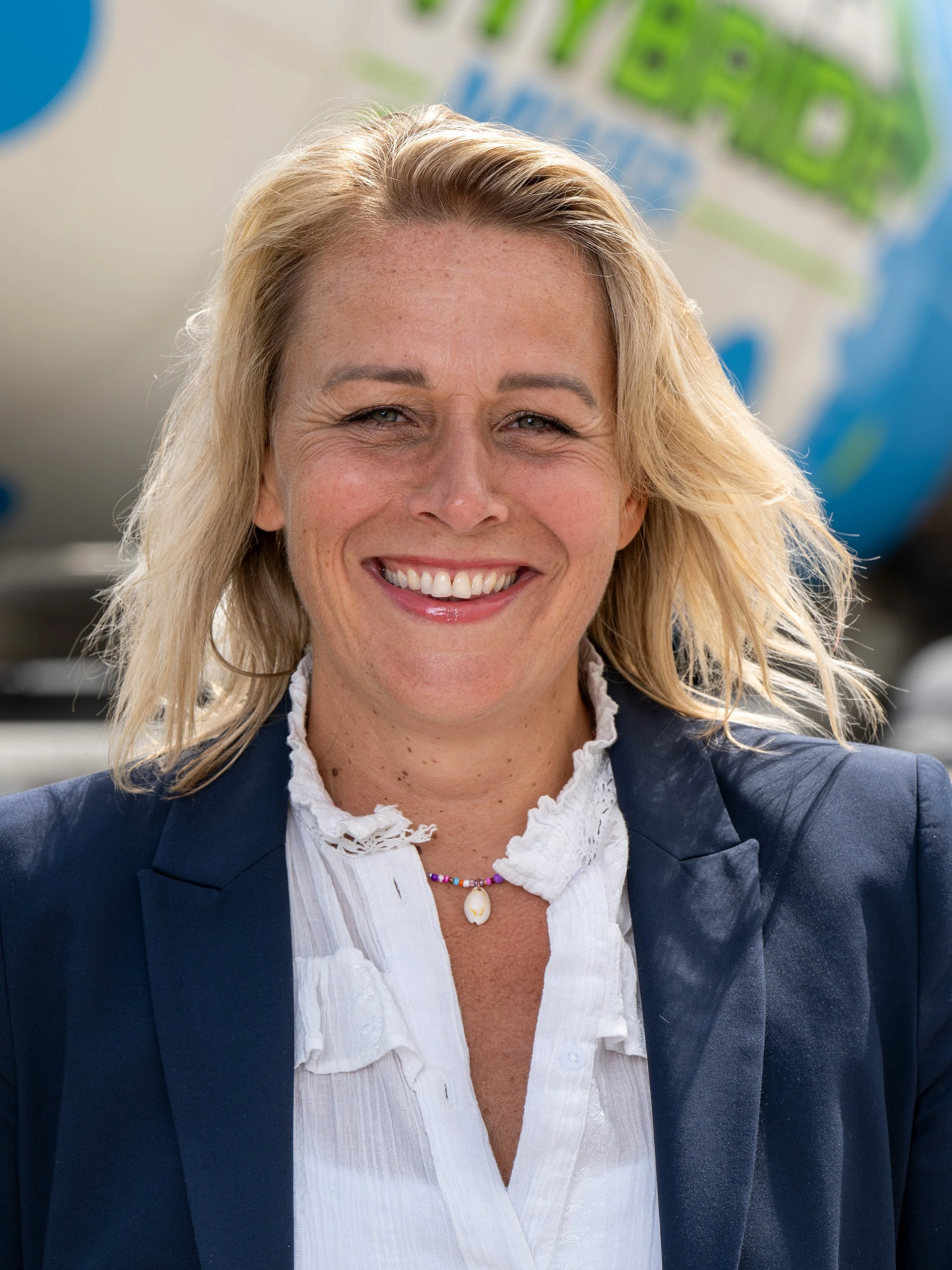 Een lachende vrouw met blond haar, gekleed in een blauwe jas en een witte blouse, met een kleurrijke ketting, achter een blauw en groen bewegwijzerd bord.