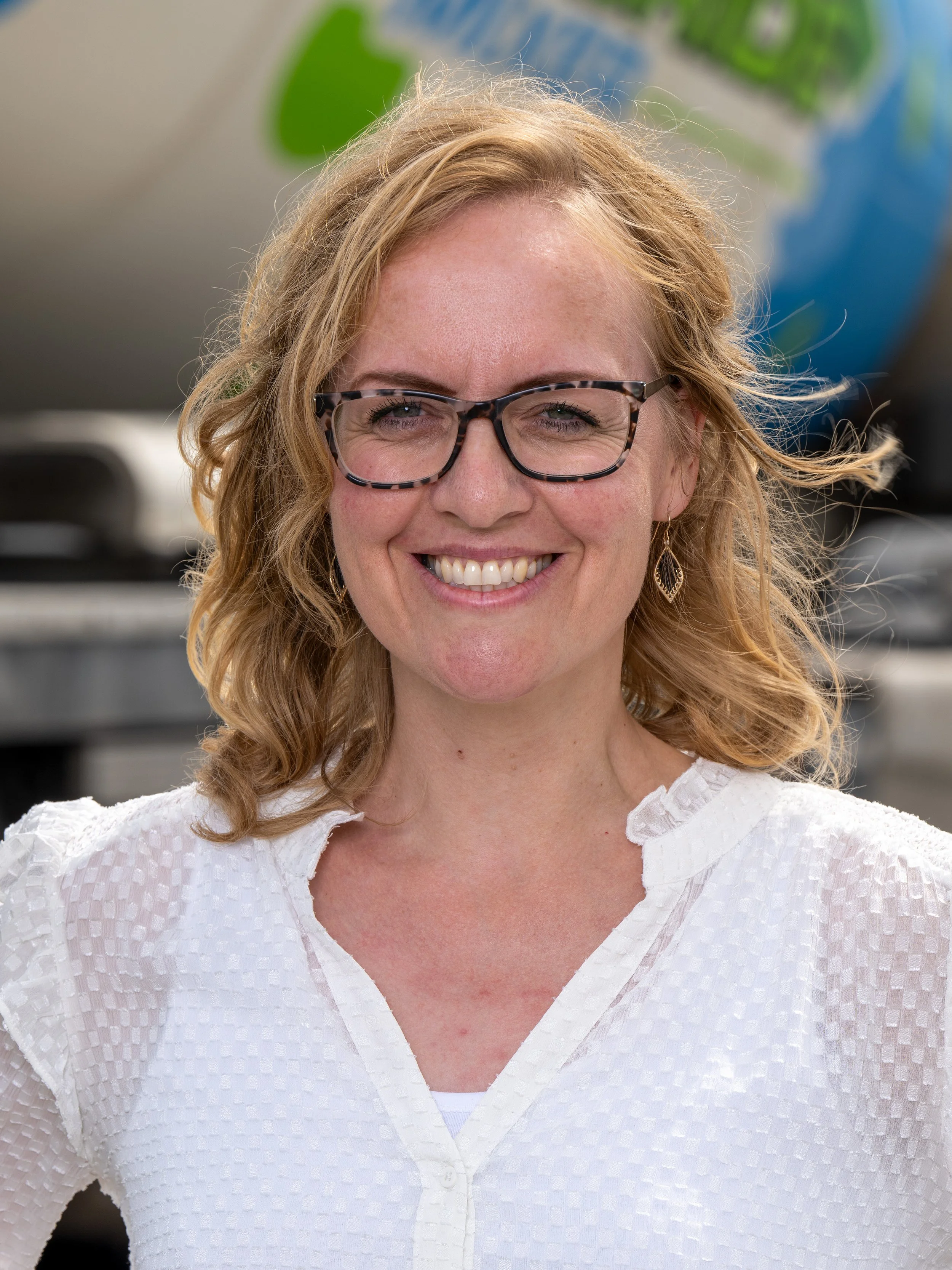 Close-up van een lachende vrouw met oranje haar, gedragen met een witte blouse en tortoise-leren brillen, met een wereldbol op de achtergrond.