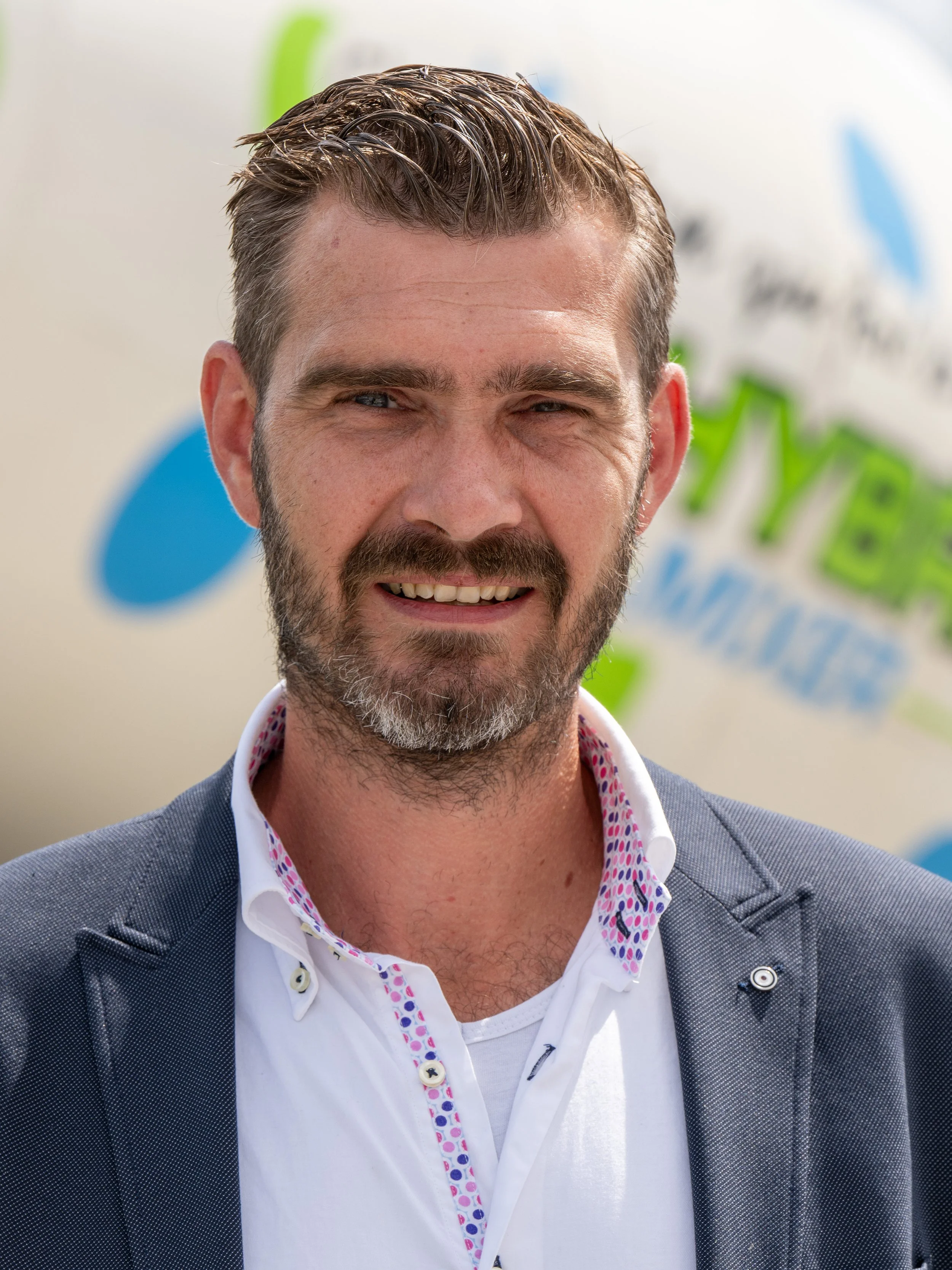 Close-up van een lachende man met een volle baard, kort haar, gekleed in een grijs jasje en een witte blouse met een kleurrijke kraag, voor een achtergrond met groene en blauwe decoraties.