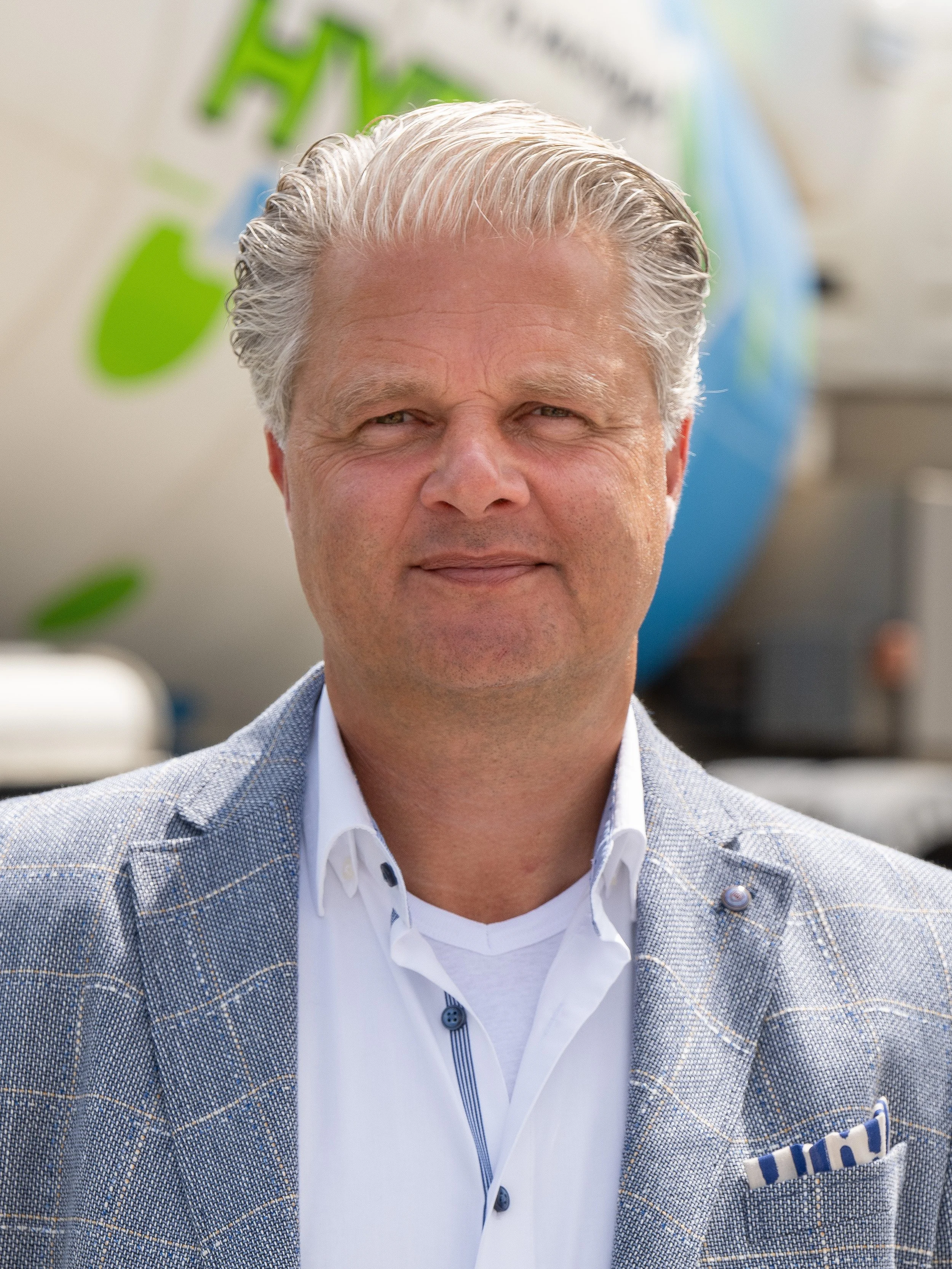 Man met lichtgrijs haar in een ruitjesblazer en een witte shirt voor een achtergrond met een globe en een groene tekst.