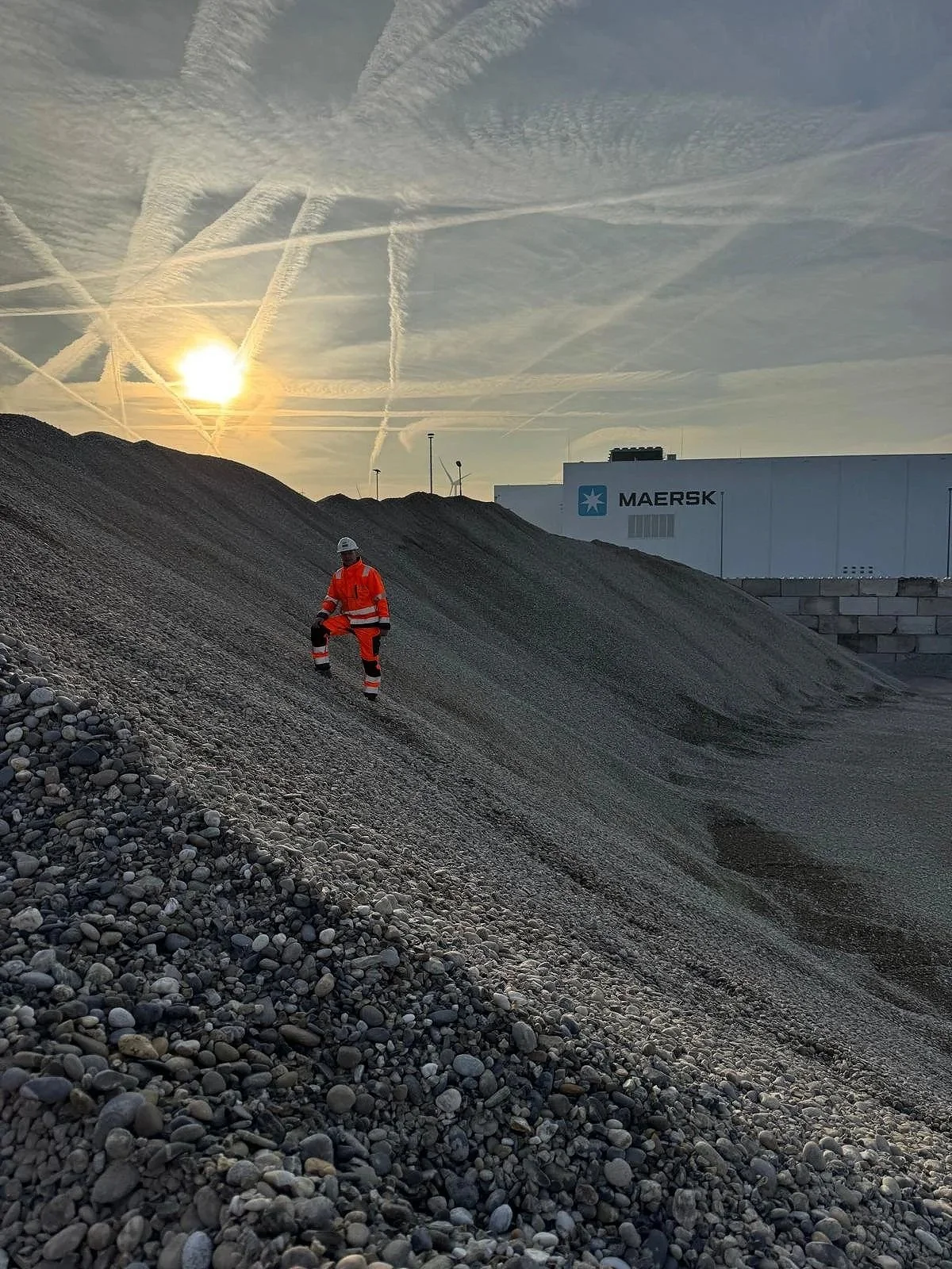 Werknemer in oranje veiligheidskleding en helm staat op een berg van grind met een achtergrond van een gebouw met de naam Maersk en een sky met veel contrails van vliegtuigen onder de zon.