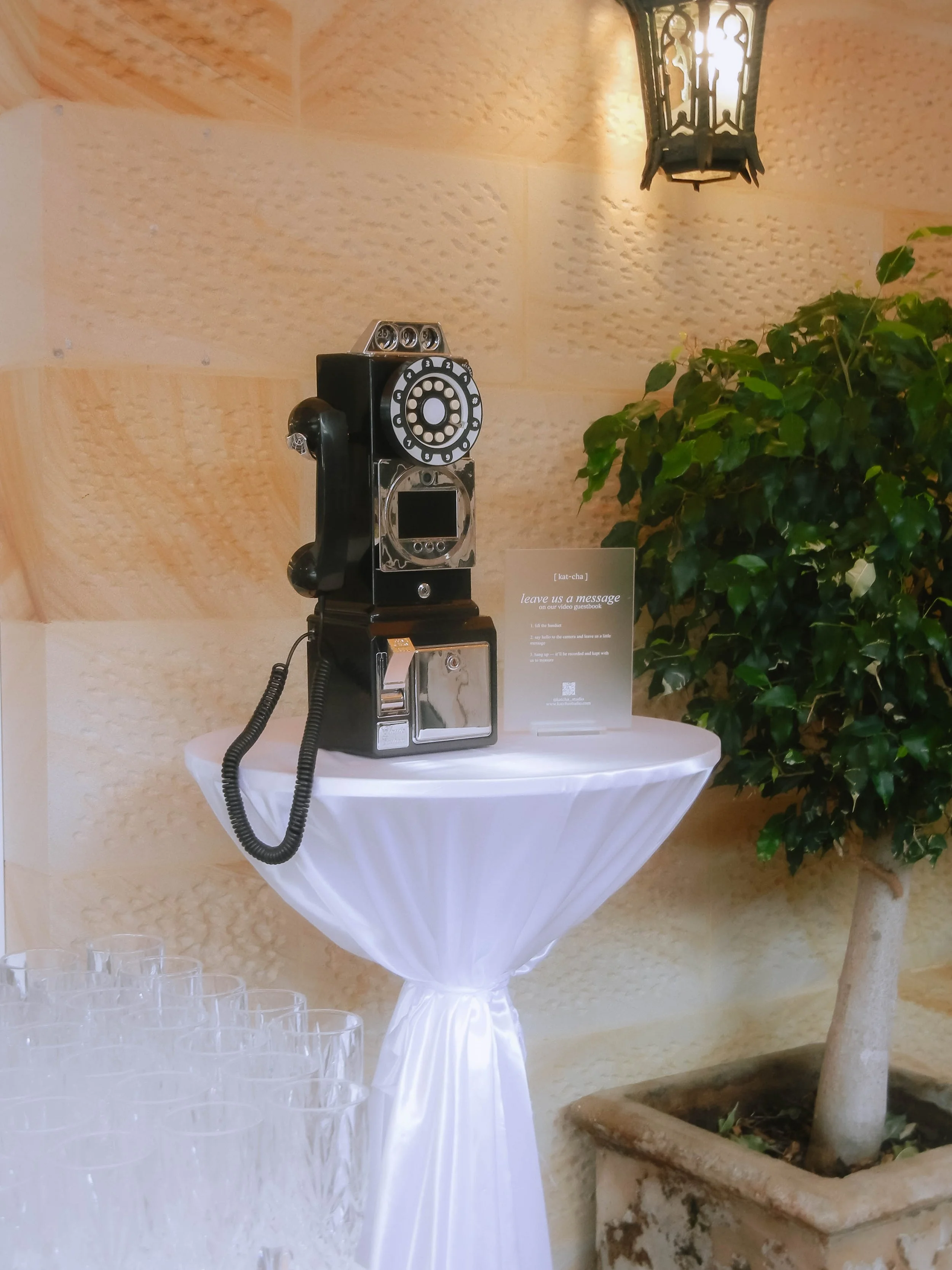A vintage black rotary phone with a silver dial and a black handset sits on a tall, round, white table covered with a white cloth. To the right, there is a potted plant with green leaves. A small sign on the table reads, 'leave us a message on our video guestbook.' In the background, there is a decorative hanging lantern with a lit bulb inside, mounted on a textured beige wall.