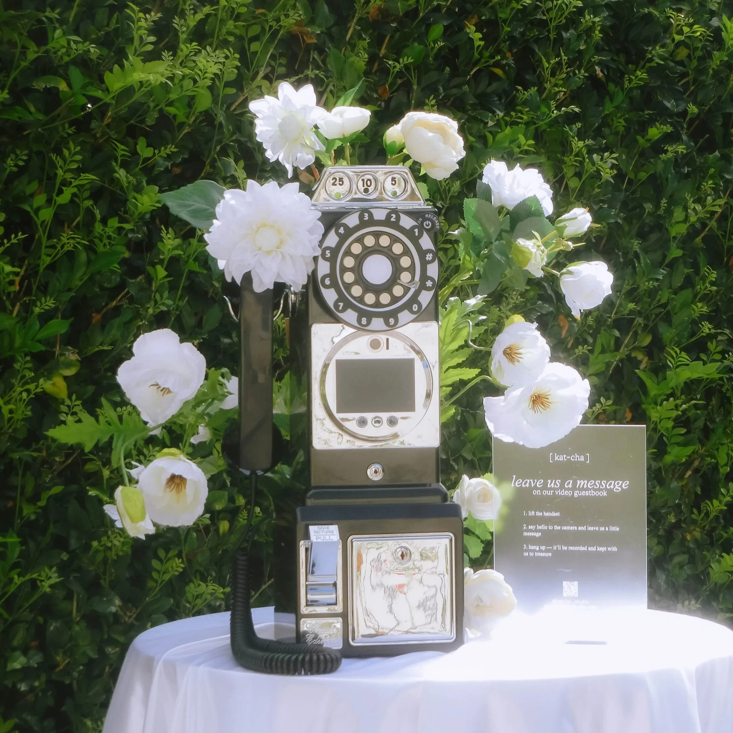 An outdoor scene with white flowers and green foliage background. A vintage style public telephone booth is placed on a white table. The booth is open, showing the rotary dial and a handset connected by a coiled cord.