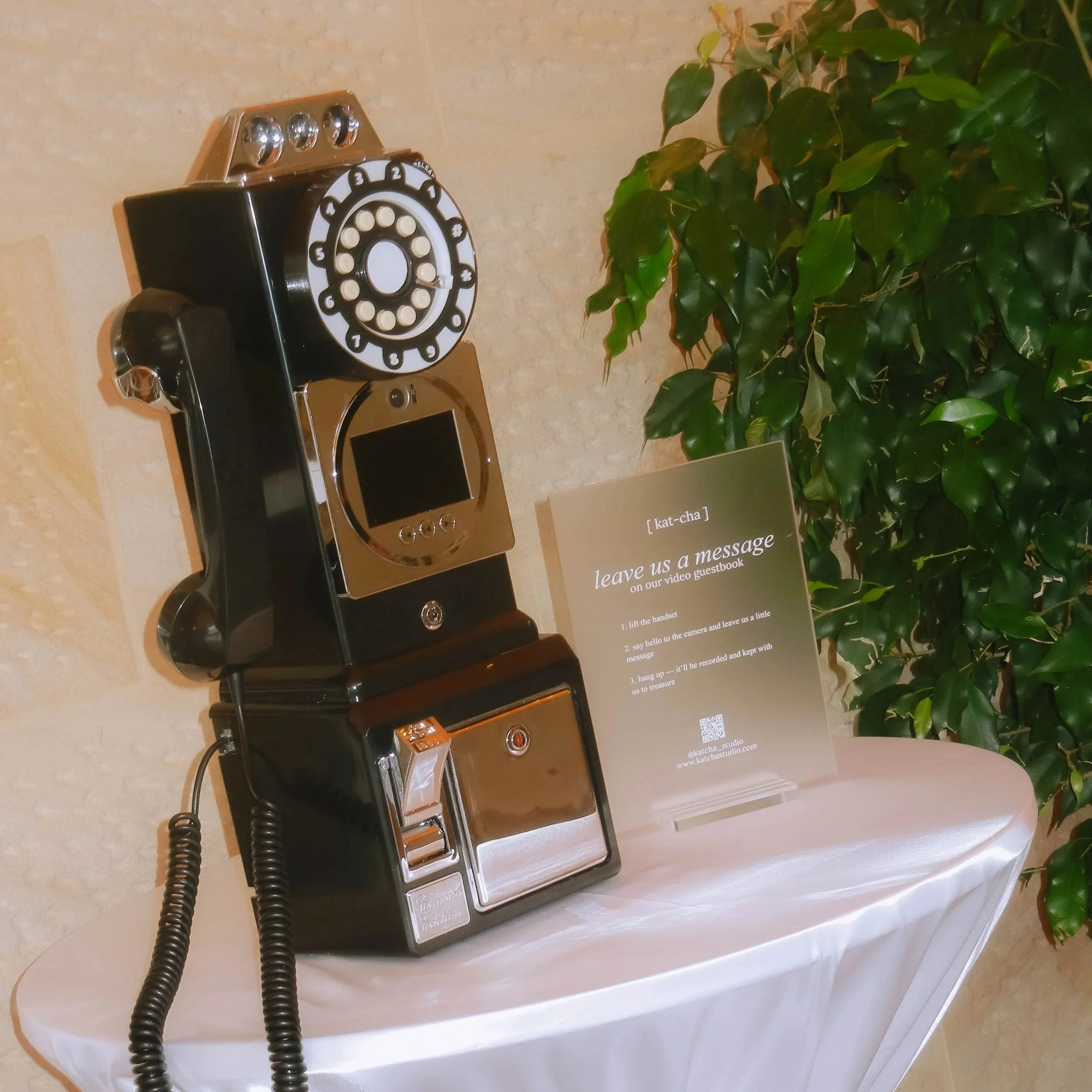 Vintage black rotary telephone with a circular dial, placed on a white tablecloth, next to a green leafy plant and a sign with instructions for leaving a message.