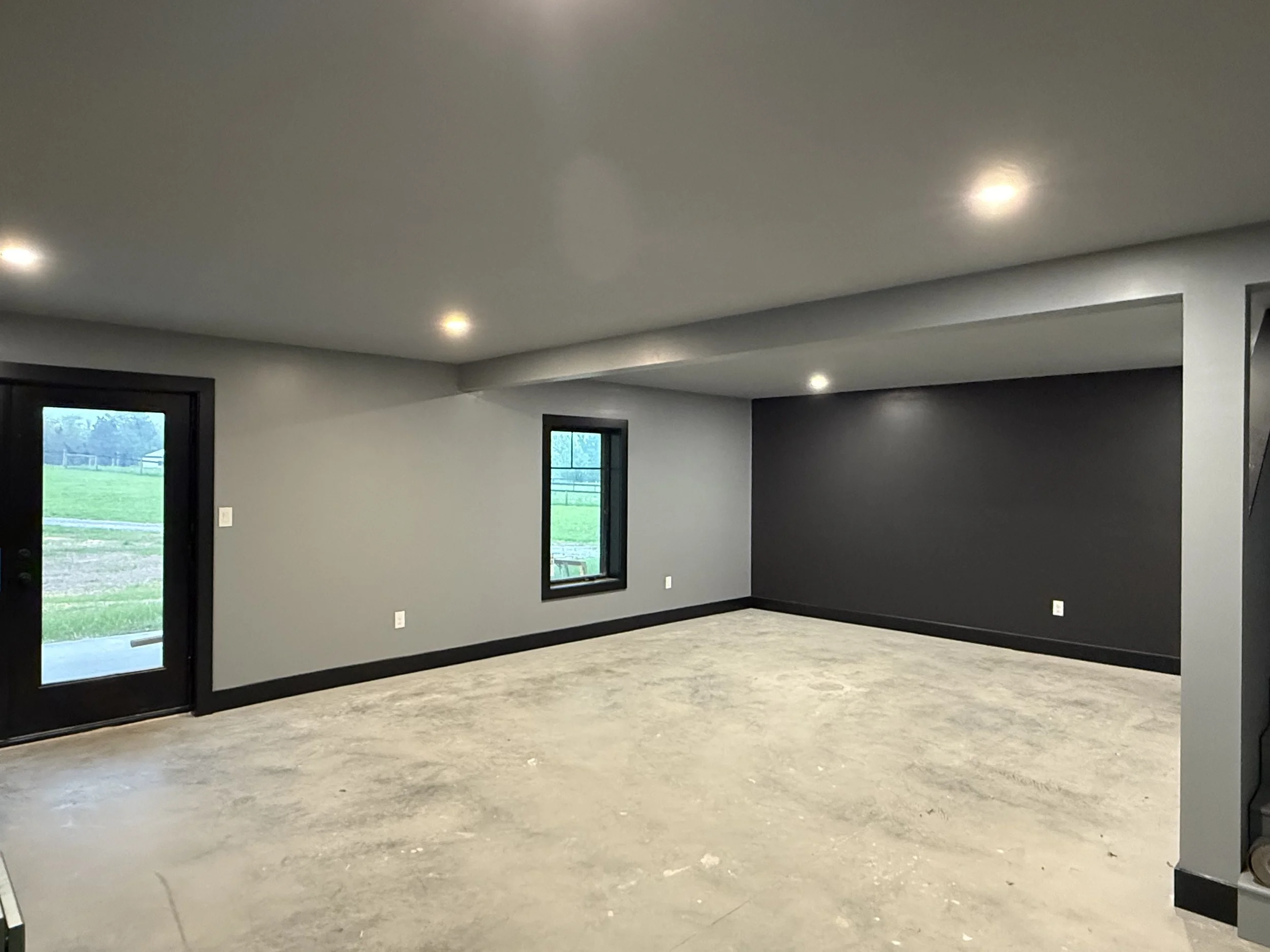 An empty room with unfinished flooring, white walls, black trims, and a dark accent wall. There are two small windows with black frames and a door with a glass panel, revealing a green outdoor landscape. Recessed ceiling lights are installed.