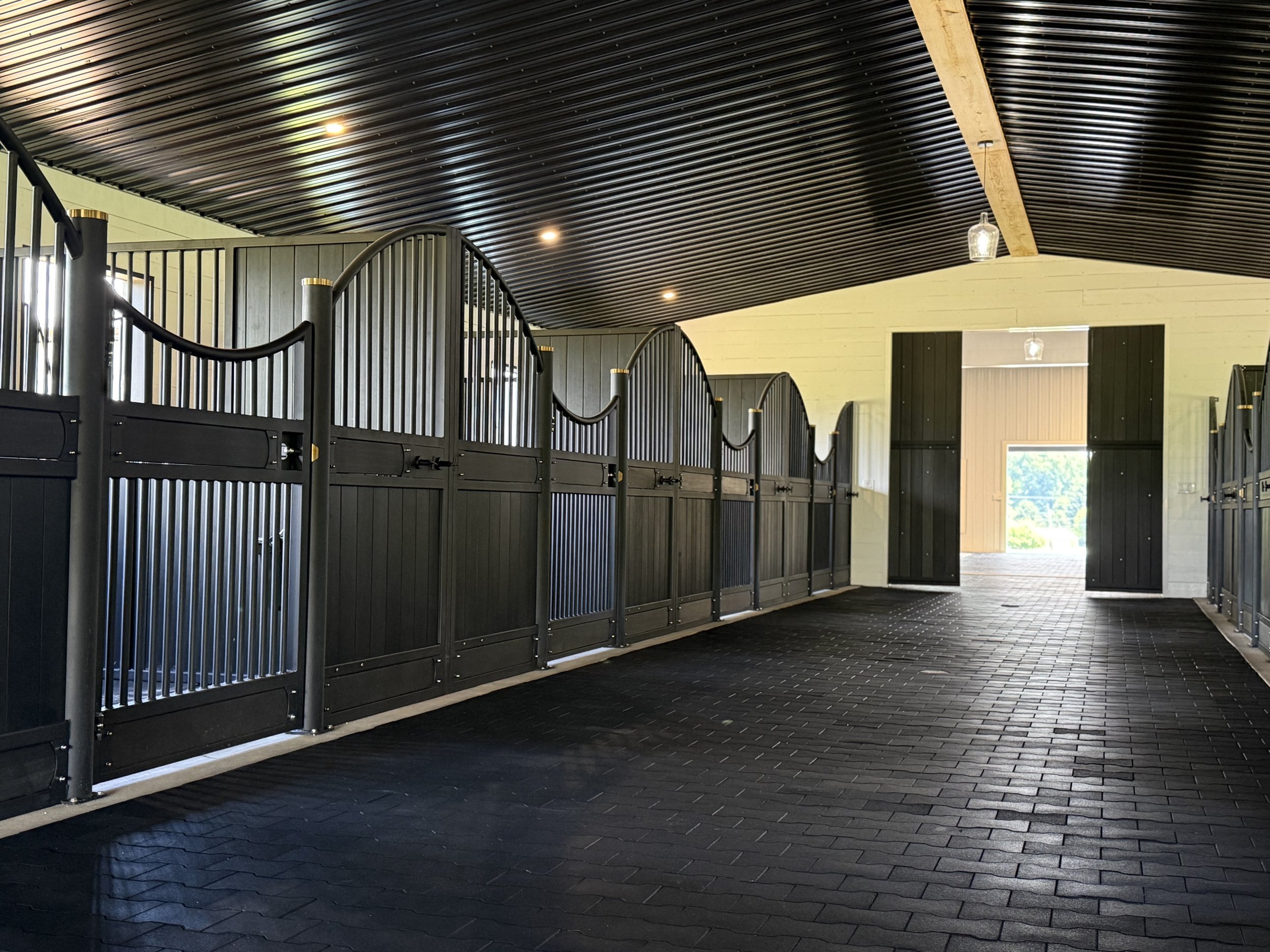 Indoor show barn with black partitioned stalls, black flooring, and a wood-paneled wall with open door leading outside.