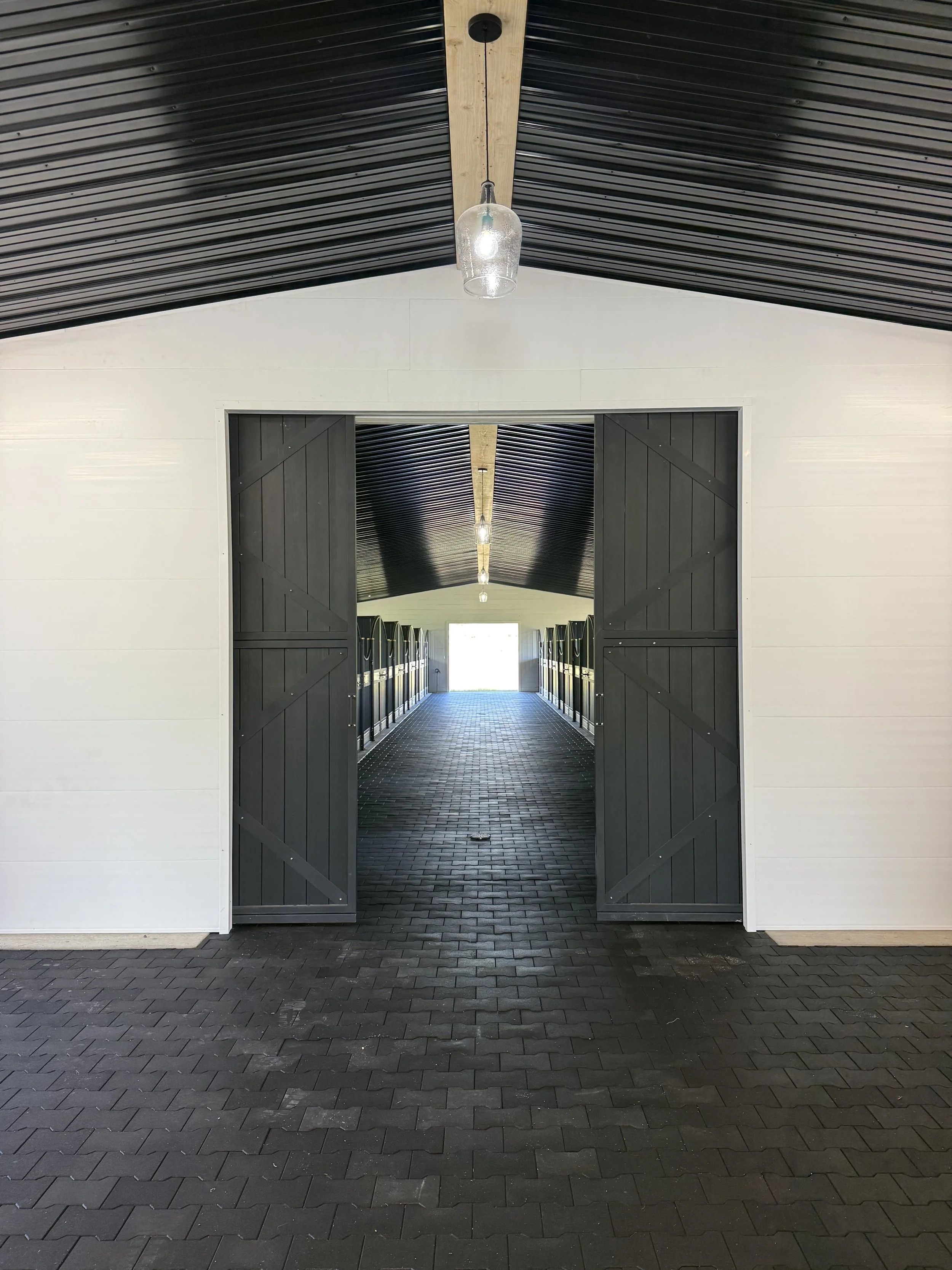 Open barn door revealing a long corridor with black lockers on both sides and hanging lights on the ceiling, leading to an open doorway with bright natural light outside.