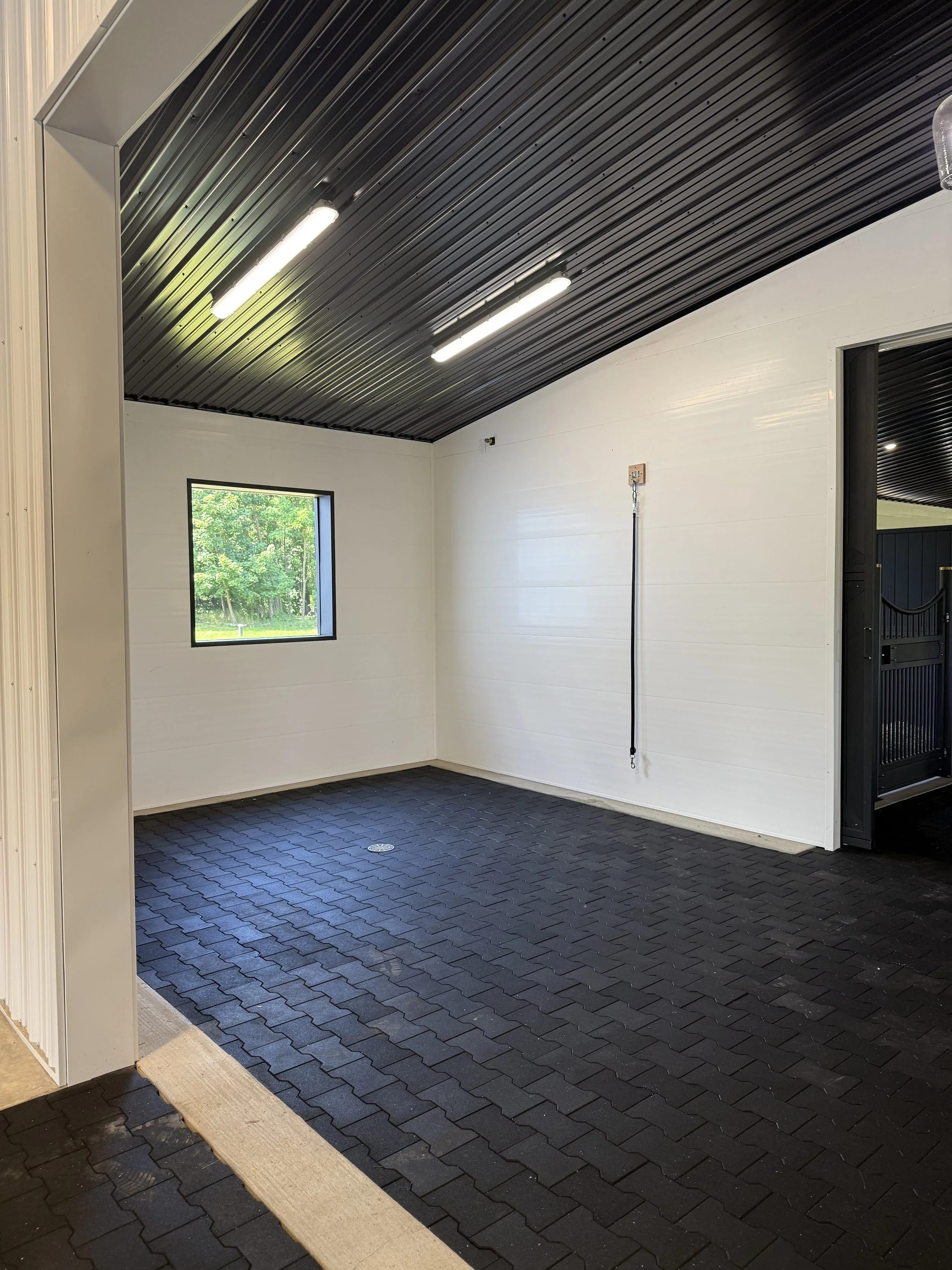 The image shows the interior of a garage with a black rubber paver floor, a white wall with a small window, fluorescent ceiling lights, and a black garage door.