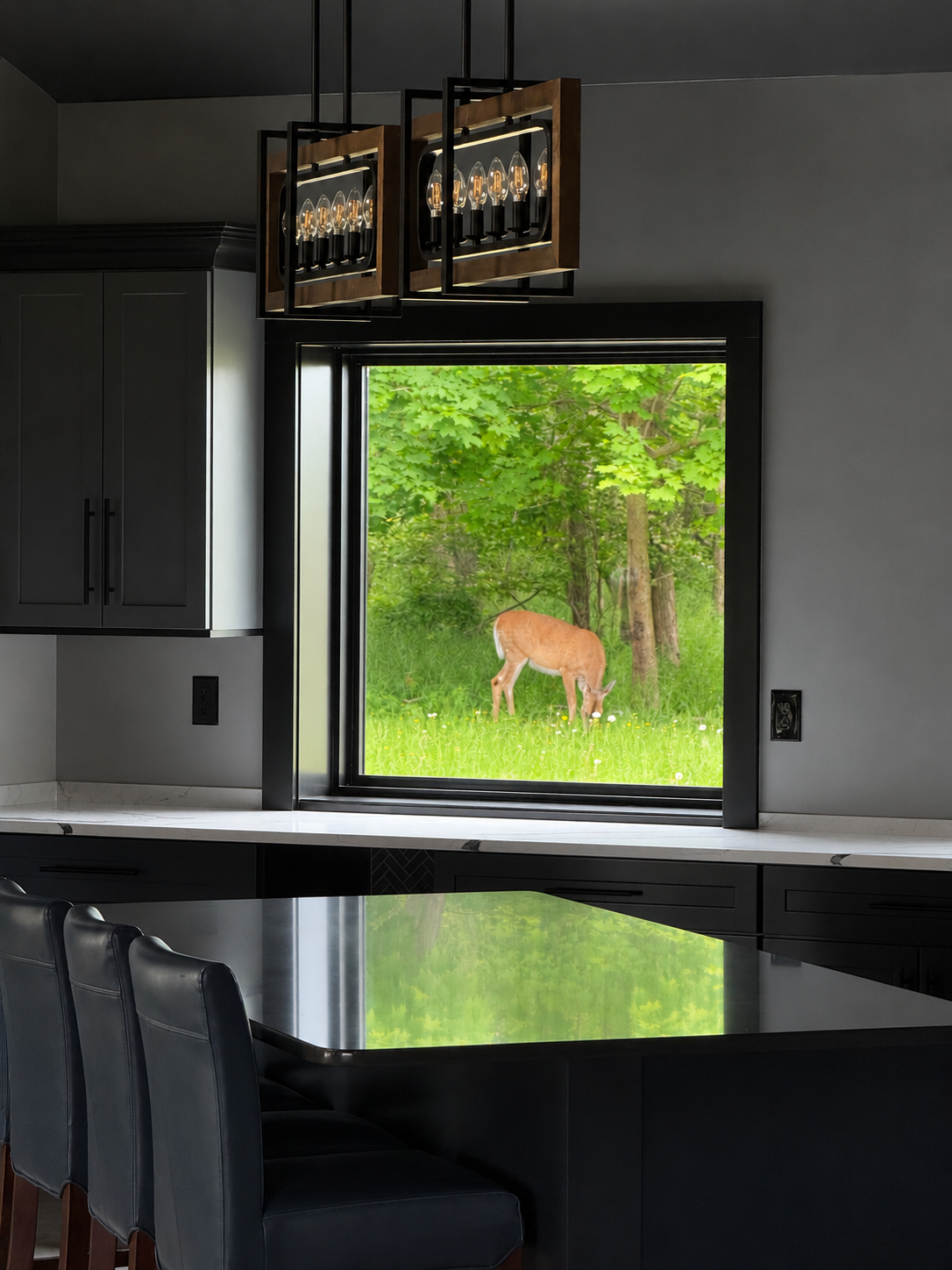 Modern kitchen interior with black cabinets, a marble countertop, black dining chairs, and a large window showing a deer grazing outside surrounded by green trees and grass.