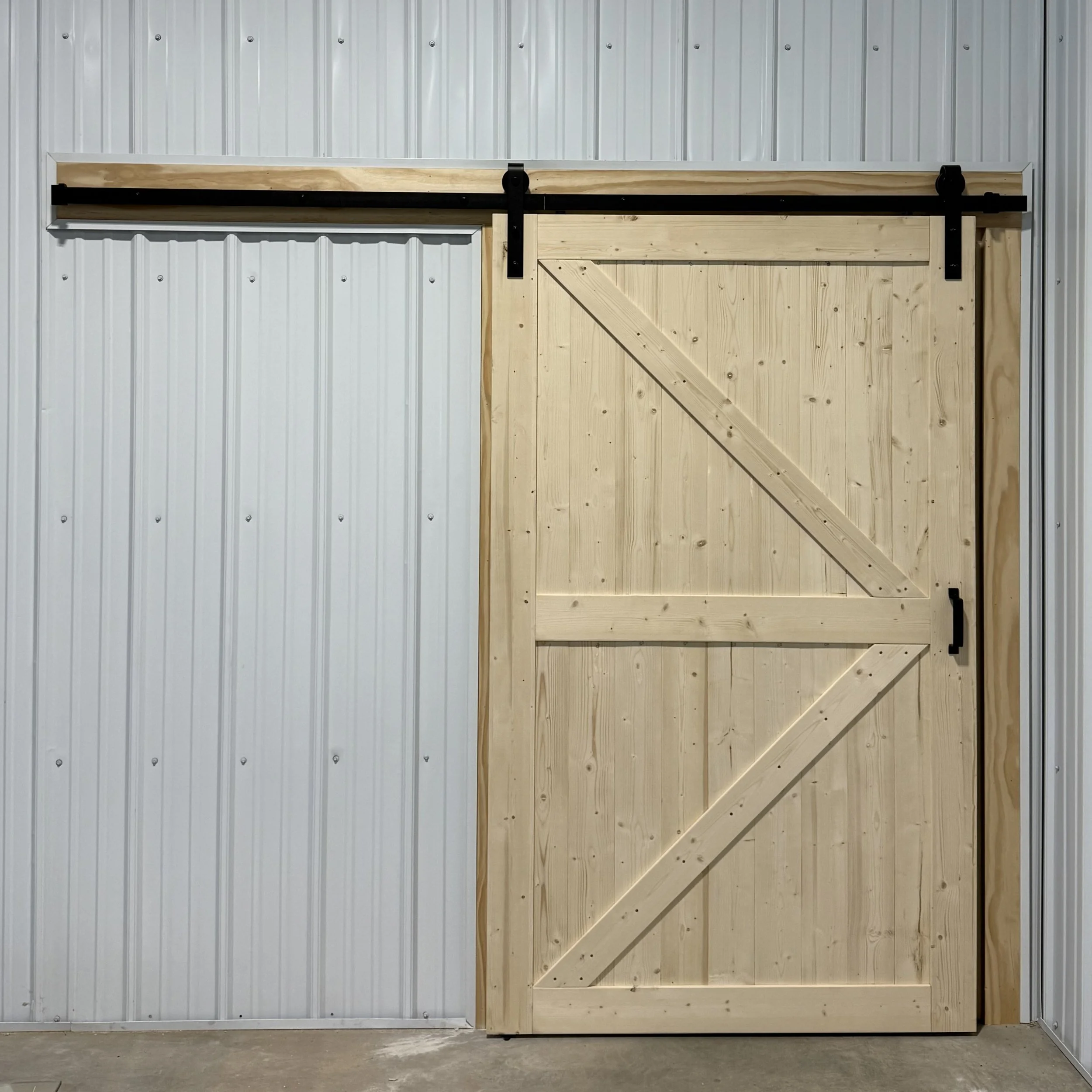 A wooden barn-style sliding door with black hardware installed on a light grey metal wall.