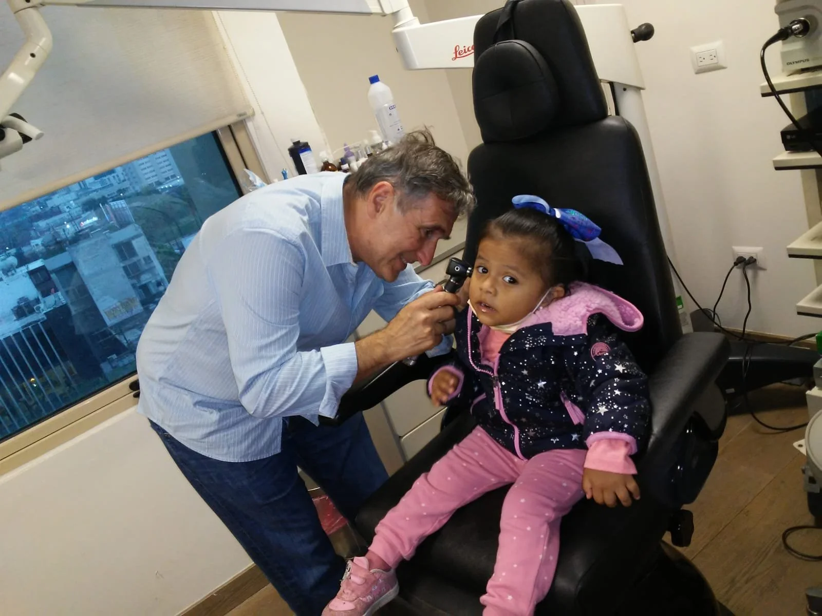 A young girl sitting in an eye examination chair at a clinic, receiving an eye test from a male doctor who is using an ophthalmoscope, with medical equipment and a window in the background.