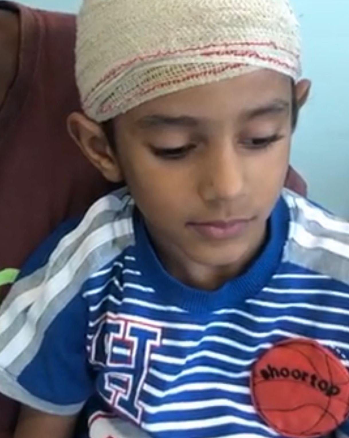Young boy wearing a blue and white striped shirt with a basketball logo, sitting with a bandage on his head.