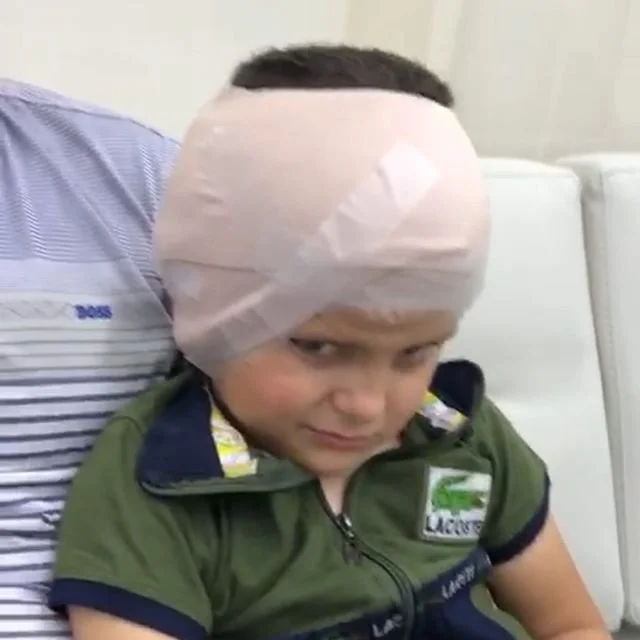 A young boy with a bandage or hat wrapped around his head, sitting on a white surface and looking up with a slight smile.