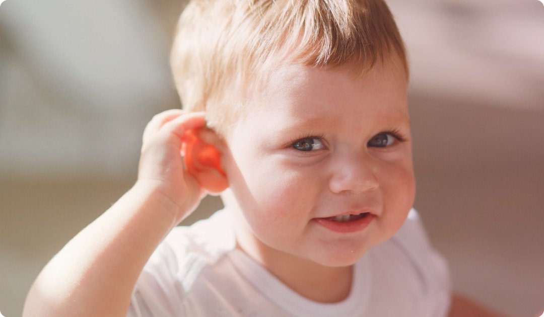 Close-up of a young boy with red hair and blue eyes smiling and cupping his hand behind his ear.