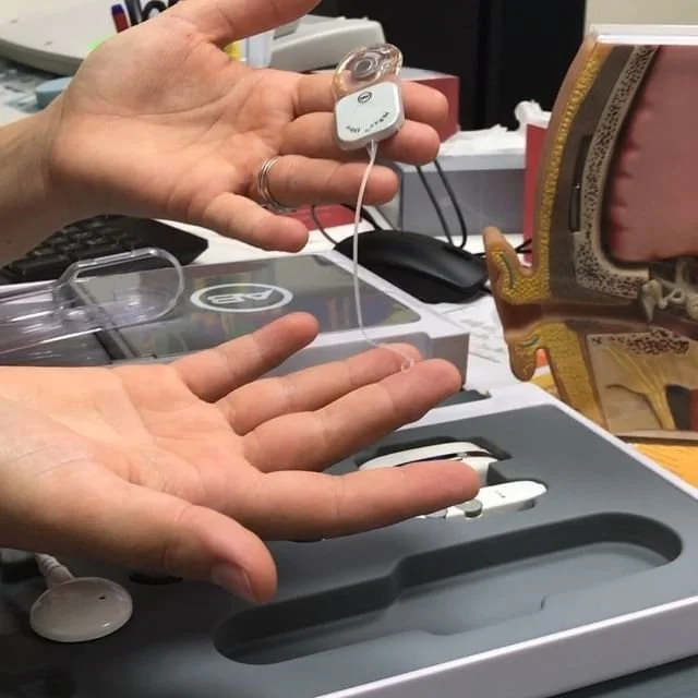 Person holding a digital thermometer above an open box with a white electronic hearing aid inside, on a desk with various objects.