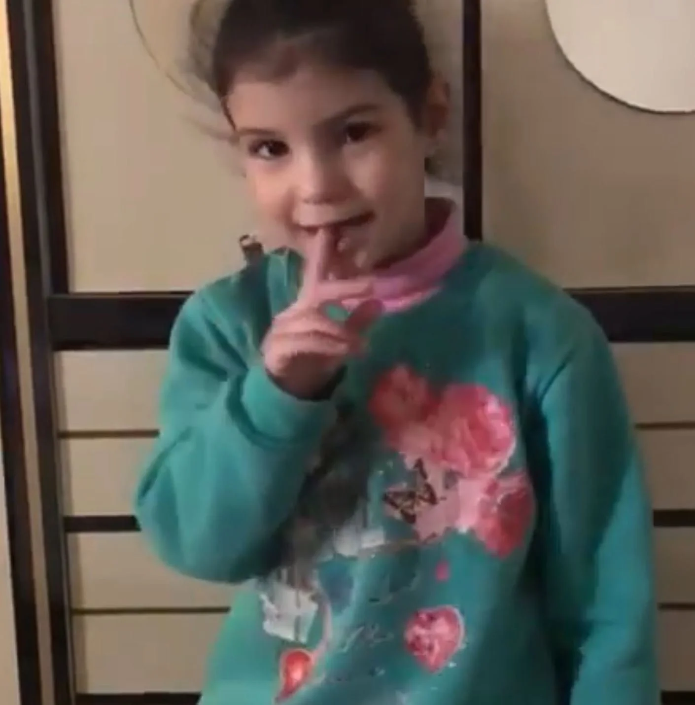 A young girl with dark hair is making a finger gesture called 'shh' near her lips, smiling slightly, in front of a beige background with black lines.