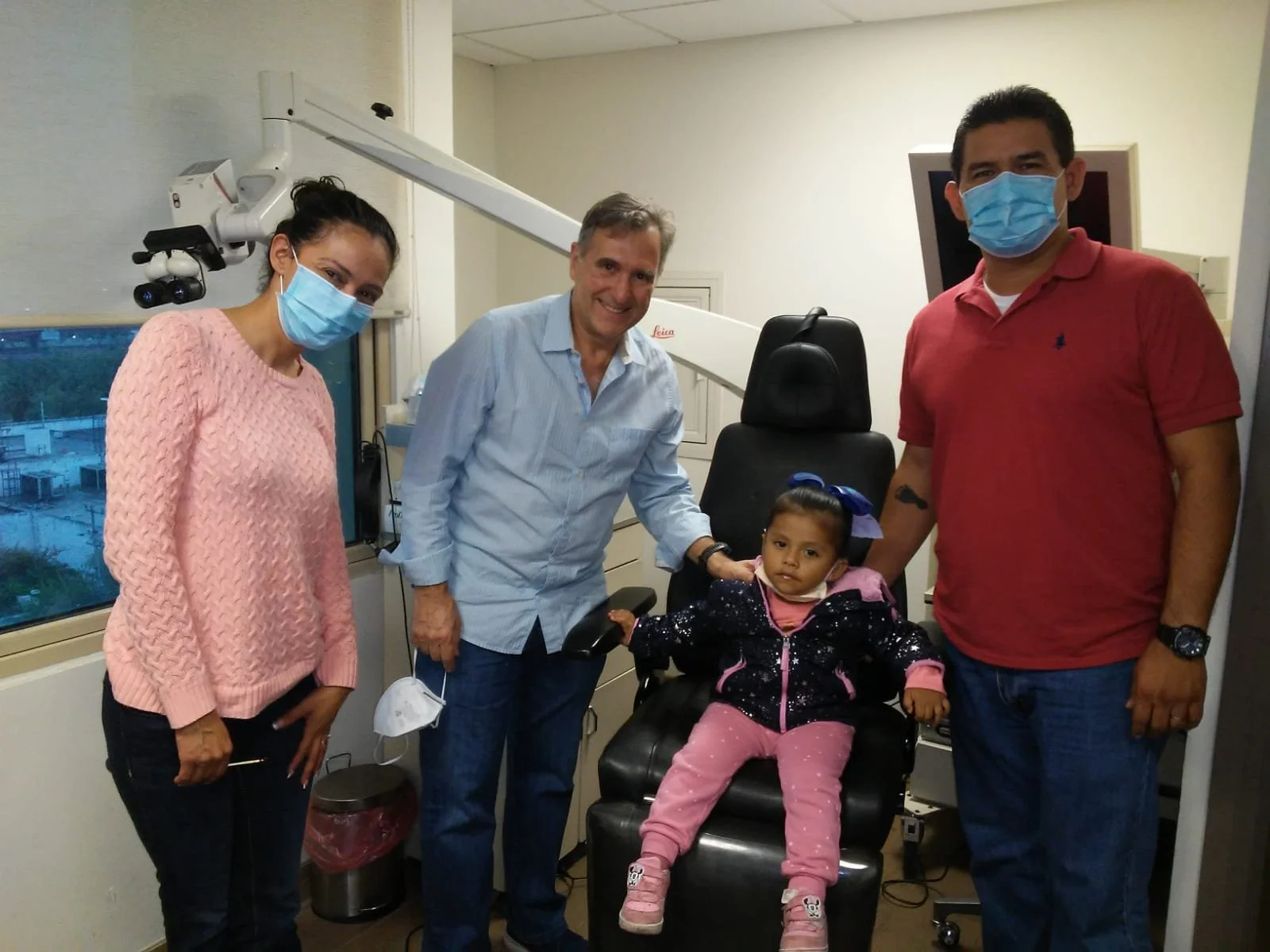 Group of four adults and one young girl in a medical examination room. The girl is sitting in a dental chair, wearing a black jacket with pink pants and pink shoes. The adults are wearing face masks. The woman on the left is in a pink sweater, the ma
