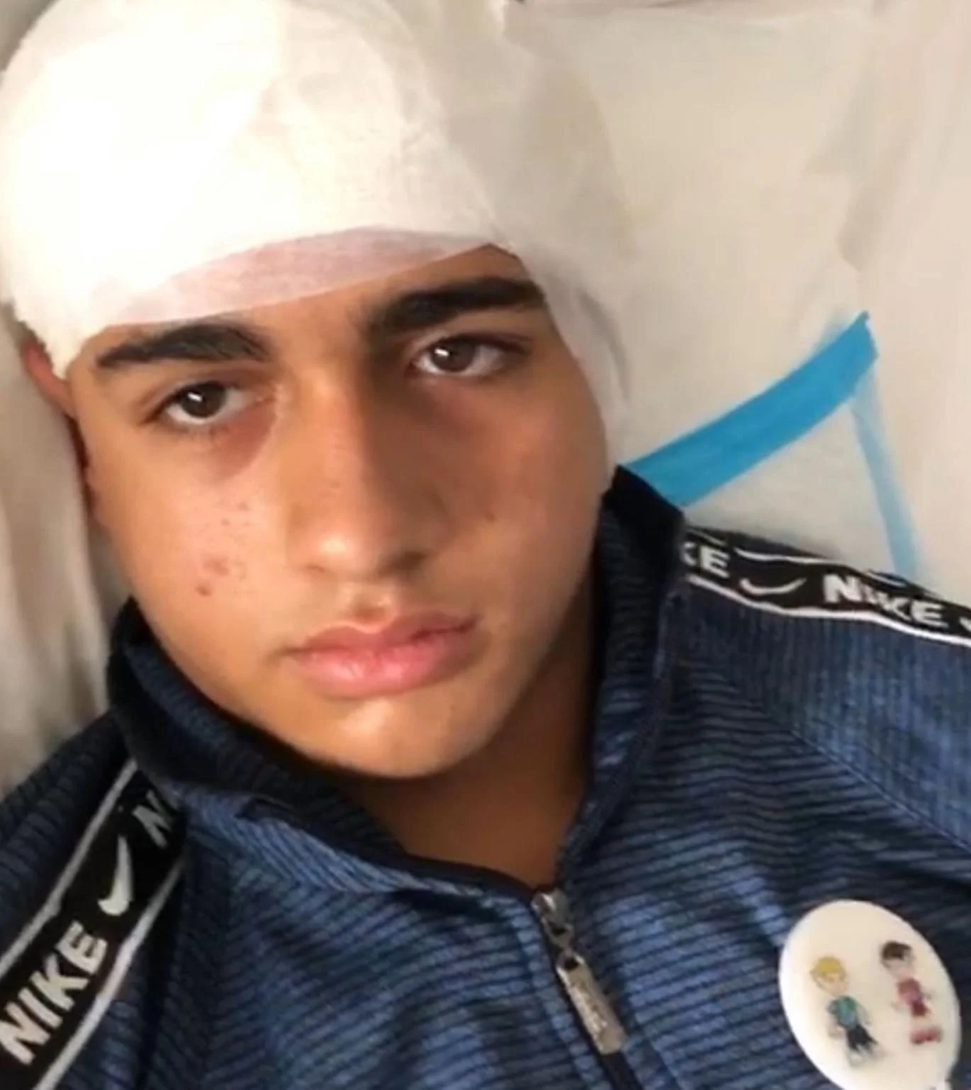 A young male with a bandage wrapped around his head, lying on a pillow, wearing a dark Nike sports jacket with a pin featuring cartoon characters on his chest.