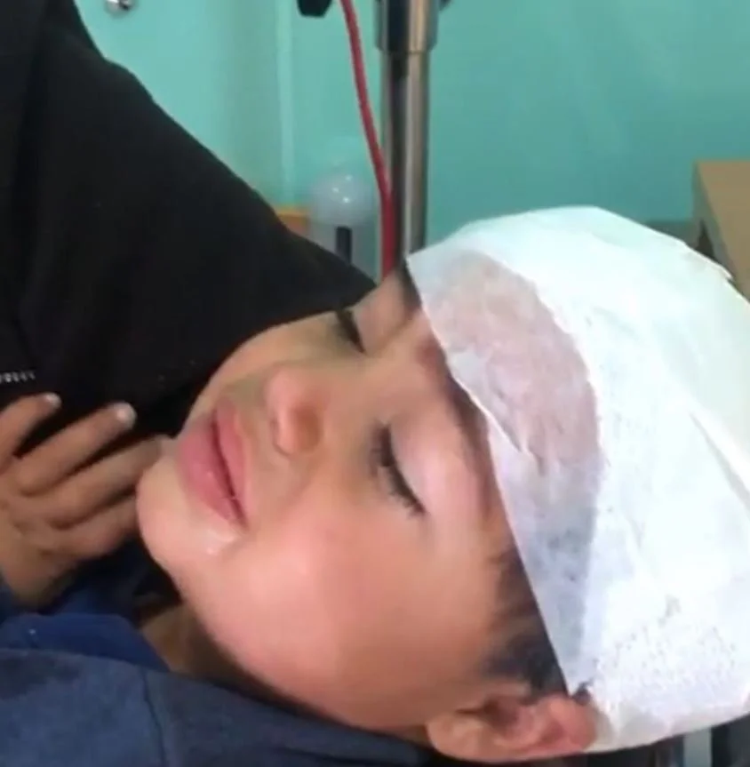 A young person is lying on a hospital bed with eyes closed, wearing a paper cap, with a bandage on their forehead, and a hand gently touching their neck. The background shows medical equipment and a greenish wall.