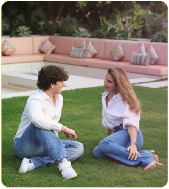 Two women sitting on grass and talking outdoors, with pink outdoor furniture and decorative pillows in the background.