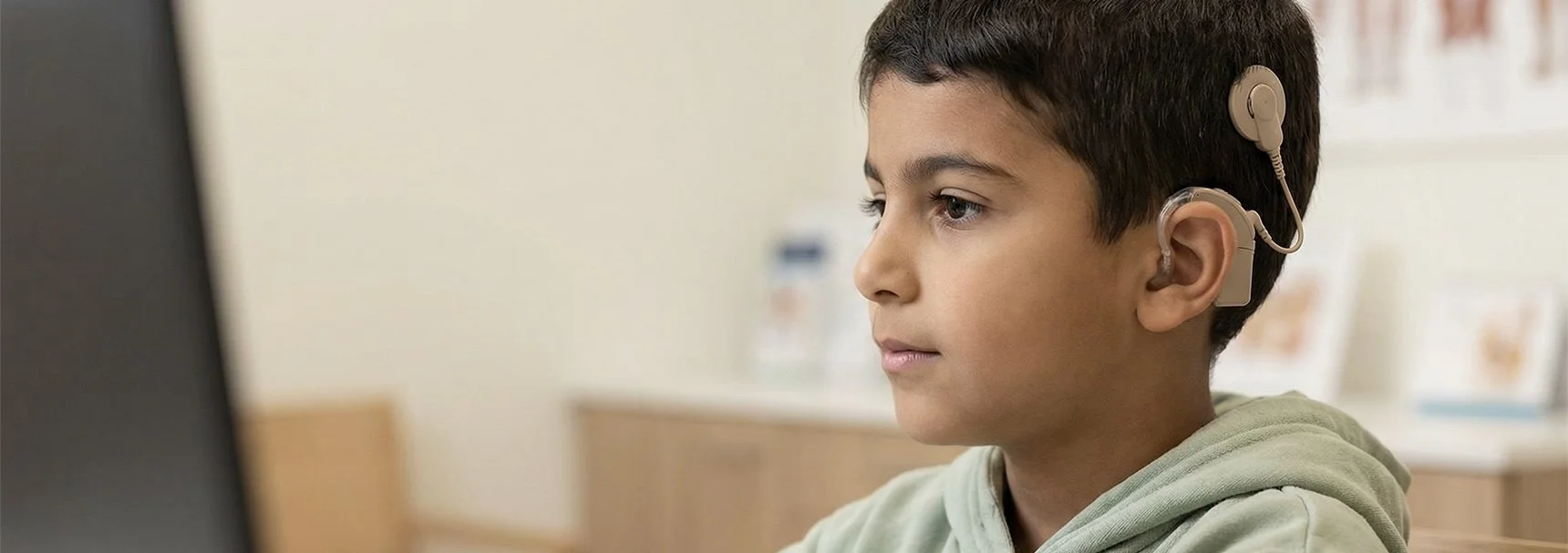 A young boy wearing a beige hearing aid sitting in front of a computer screen in a bright room.