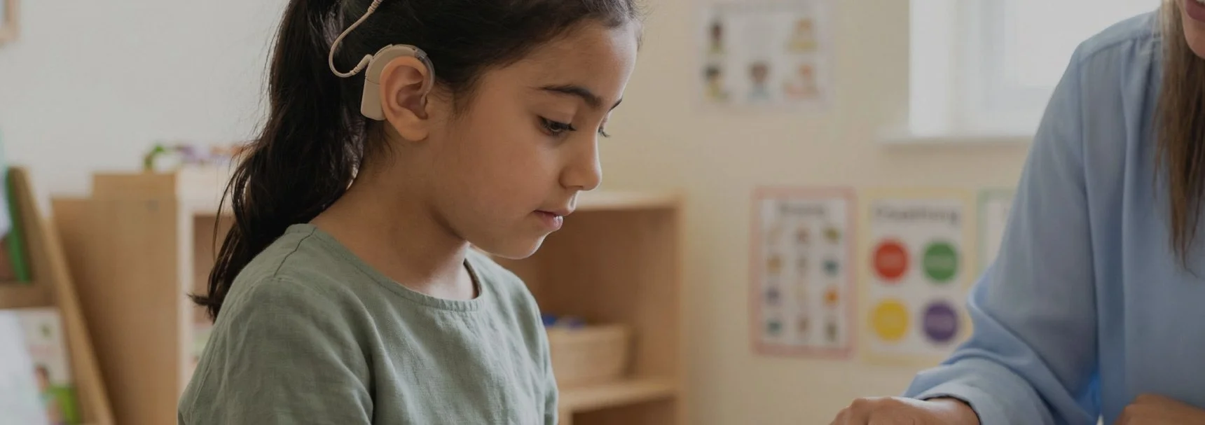 A young girl with dark hair wearing a hearing aid and headphones looks down thoughtfully in a classroom setting.