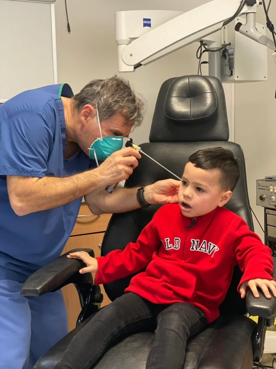 A healthcare professional wearing a blue scrubs and a face mask examines a young boy sitting in a medical chair, with medical equipment in the background.