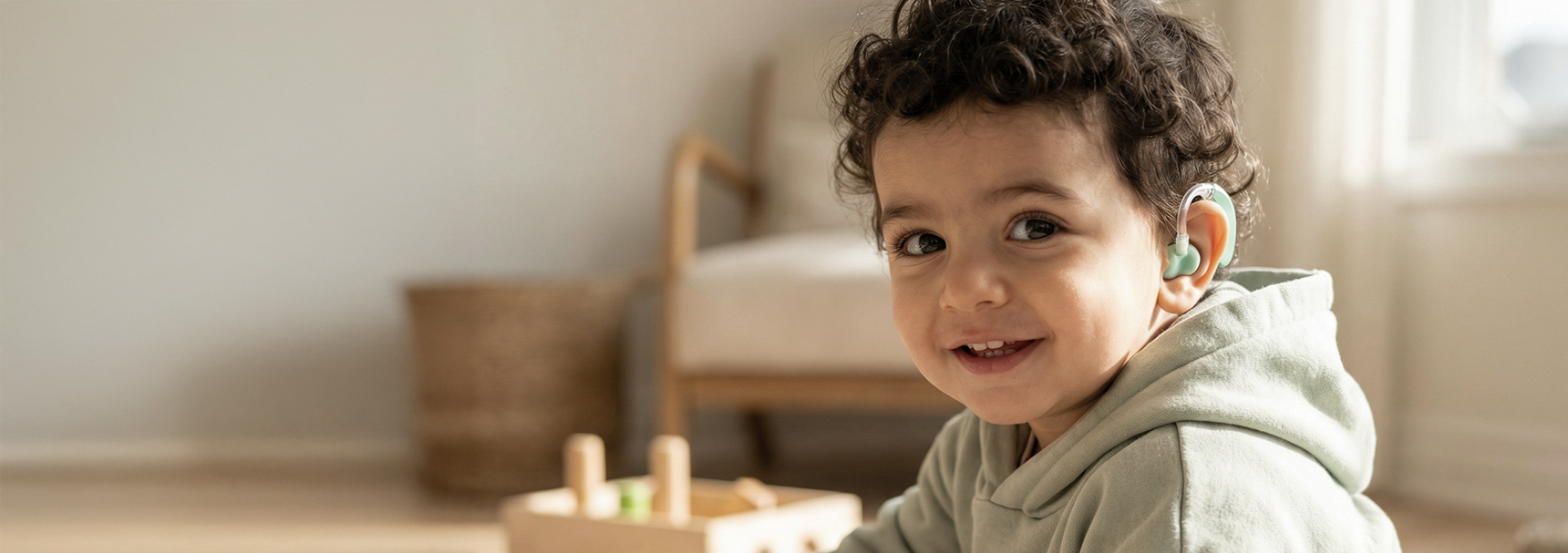 Young child with curly hair smiling and wearing a hearing aid, indoor setting with a bed and window in the background.