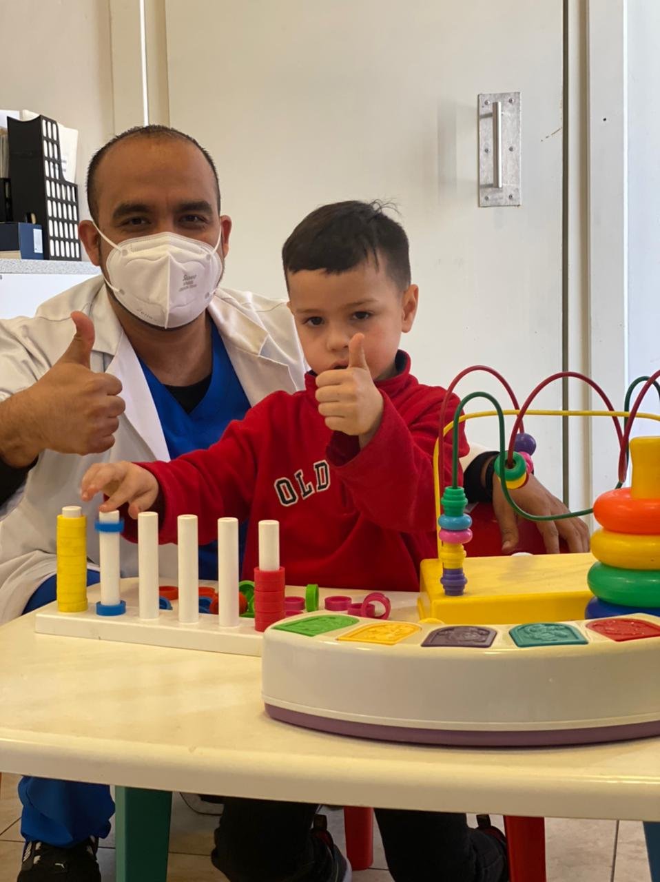 A healthcare worker and a young boy giving thumbs up at a table with colorful toys, in a healthcare setting.