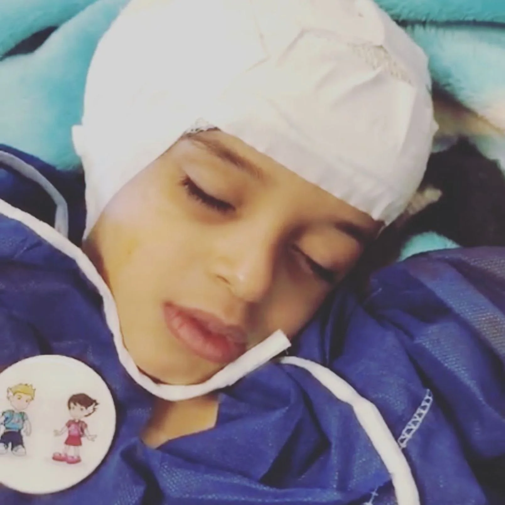 A young child with a white head band sleeping peacefully, lying on a blue blanket, with a button featuring cartoon characters on their clothing.