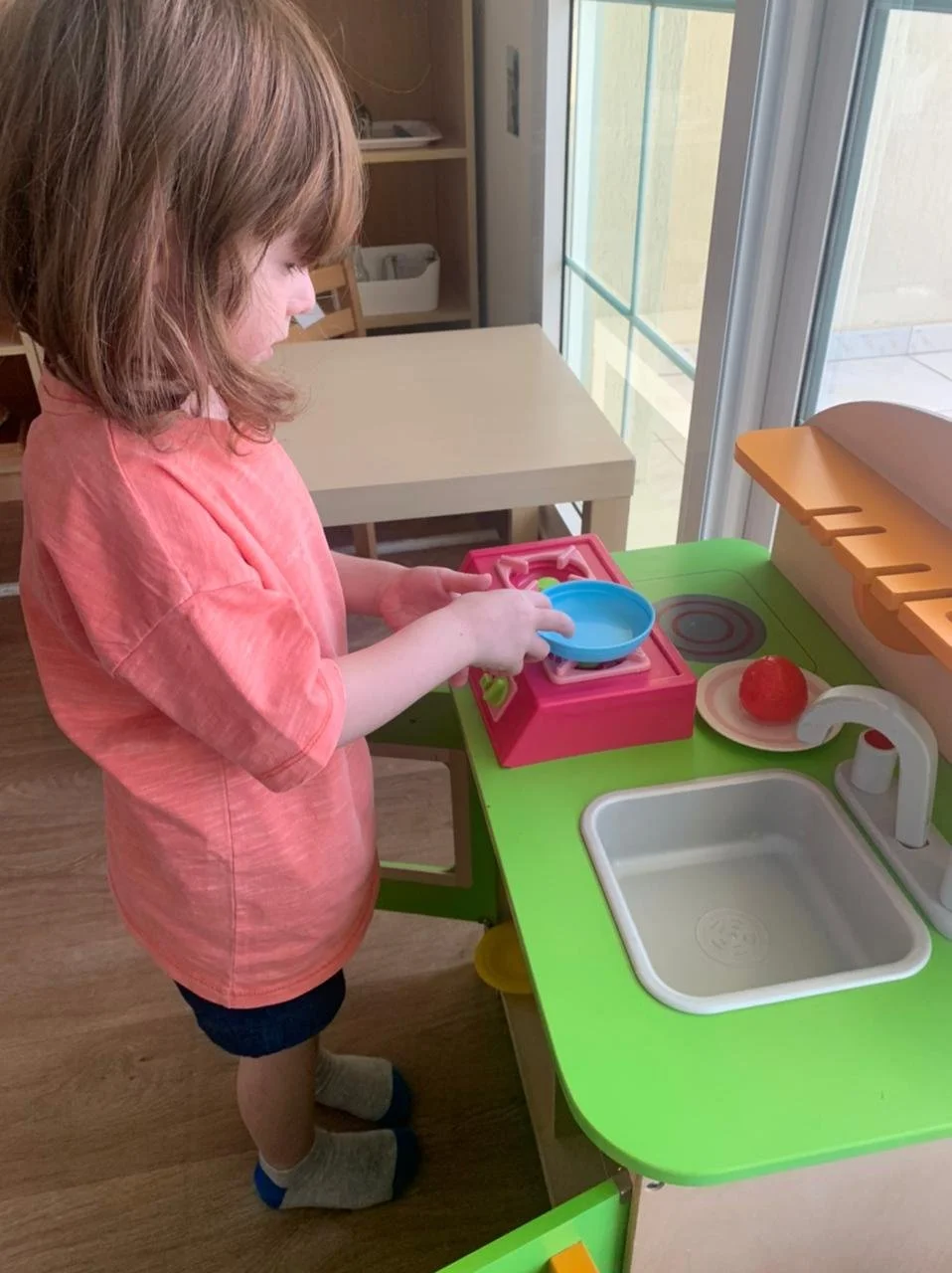A young girl with shoulder-length brown hair wearing a pink t-shirt, black shorts, and gray socks standing at a colorful play kitchen set, holding a blue toy bowl.